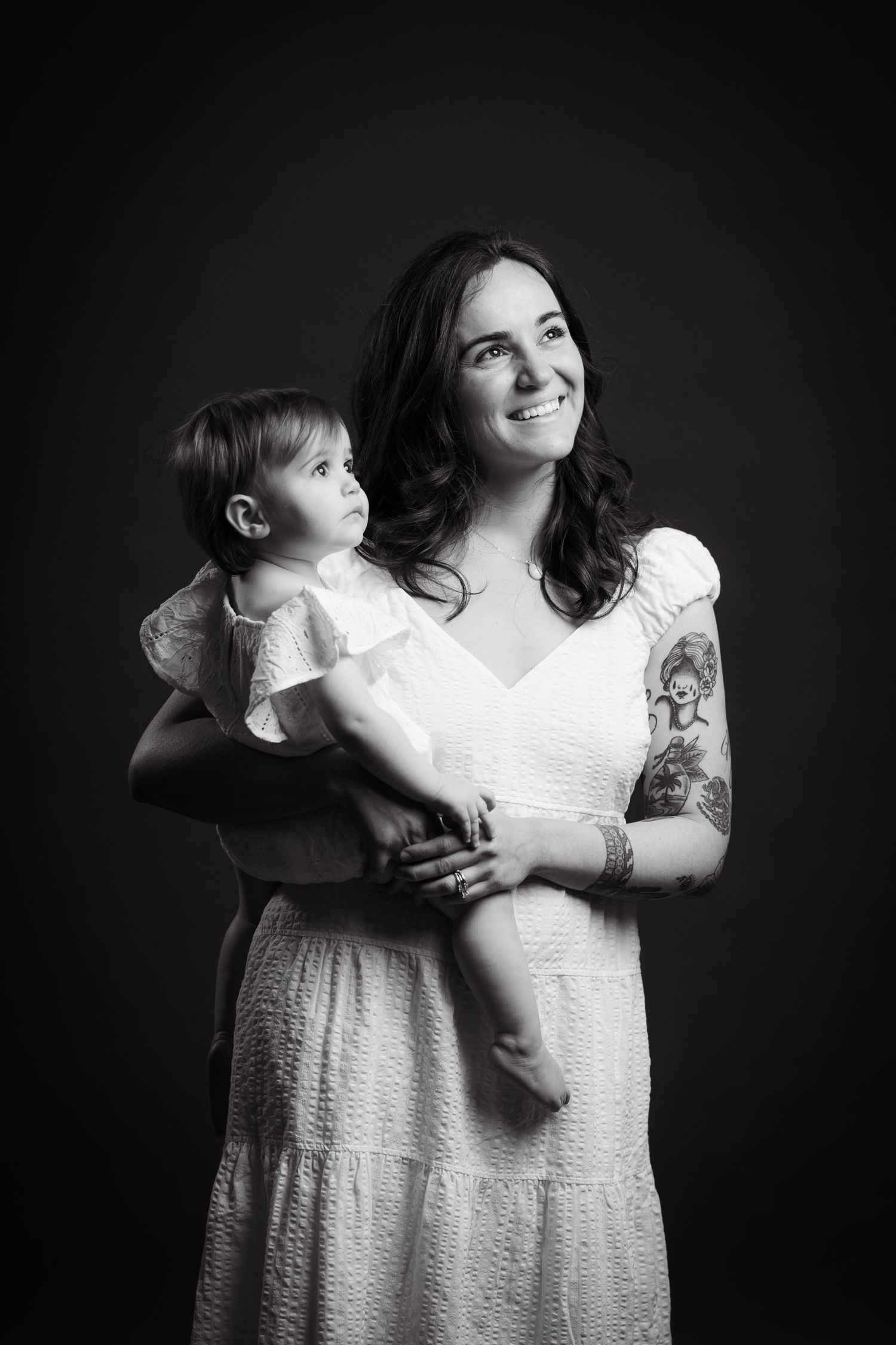 Black and white studio portrait showing mother in white dress holding small child against dark background.