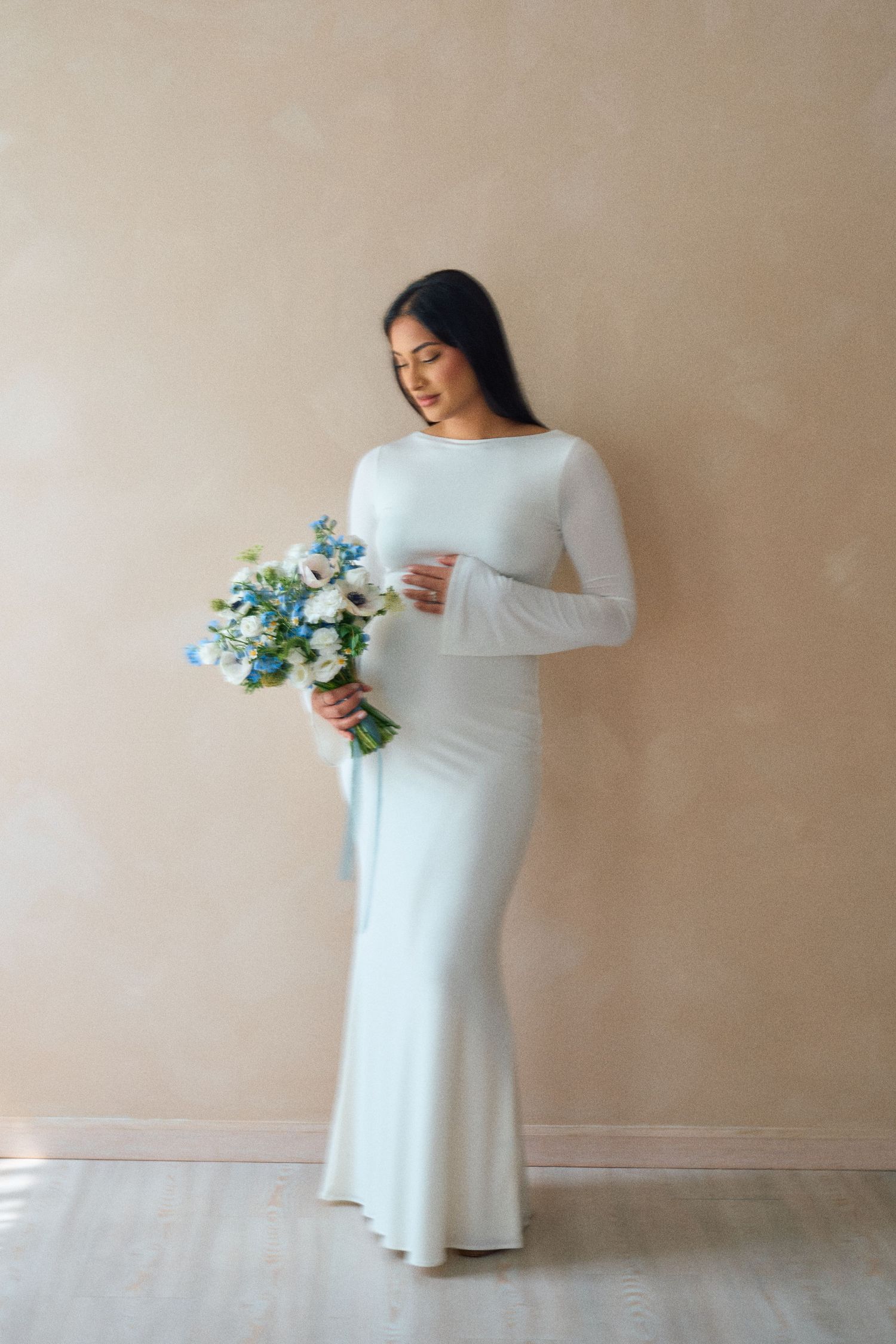A person in a long-sleeved white wedding dress holds a blue and white floral bouquet against a beige wall.