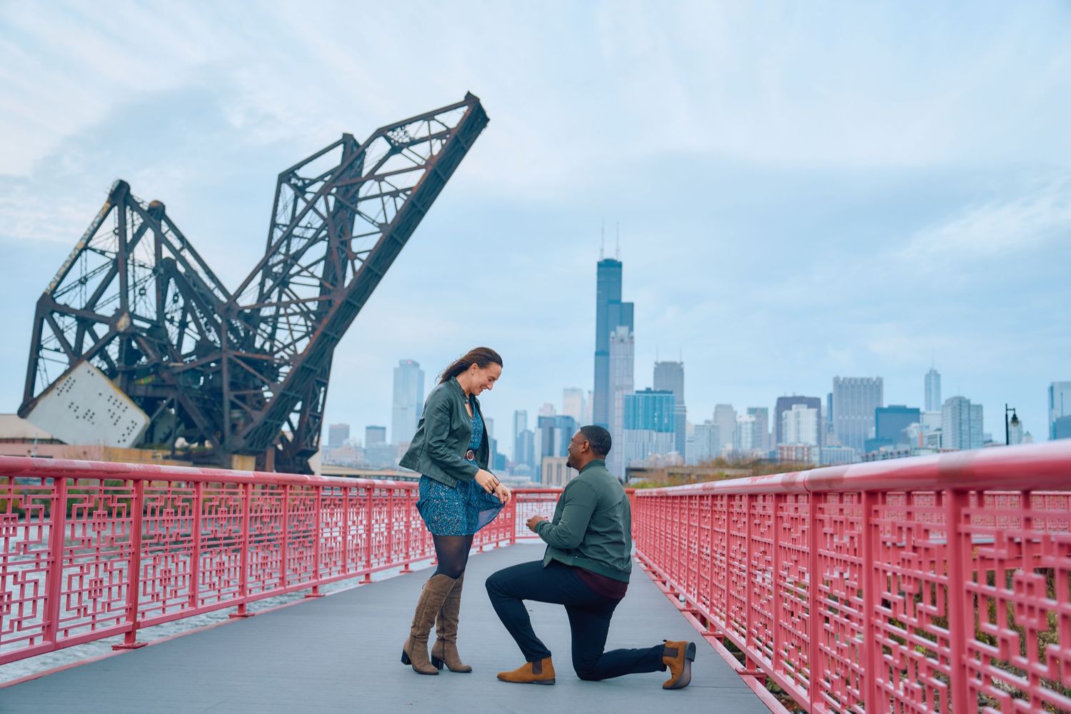 A couple getting engaged on a red bridge with the Chicago skyline and railroad drawbridge in the background.