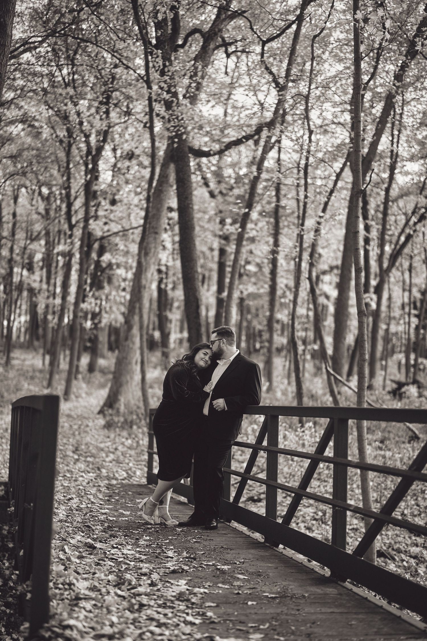 A couple embraces tenderly on a wooden bridge in an autumn forest, surrounded by bare trees in black and white.