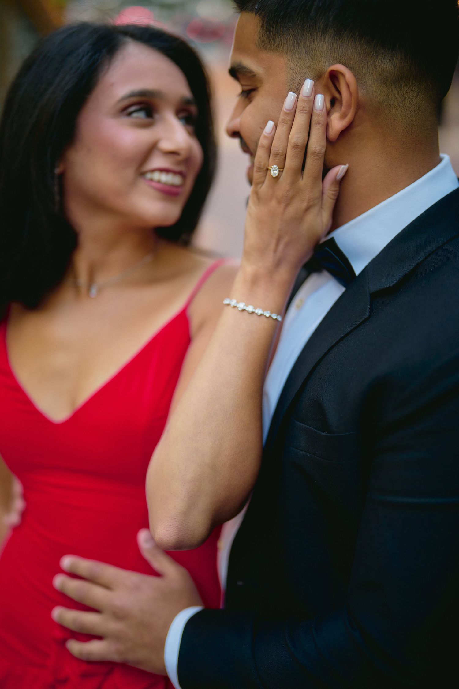 Romantic couple in formal attire sharing an intimate moment, with a red dress and black suit against a blurred background.