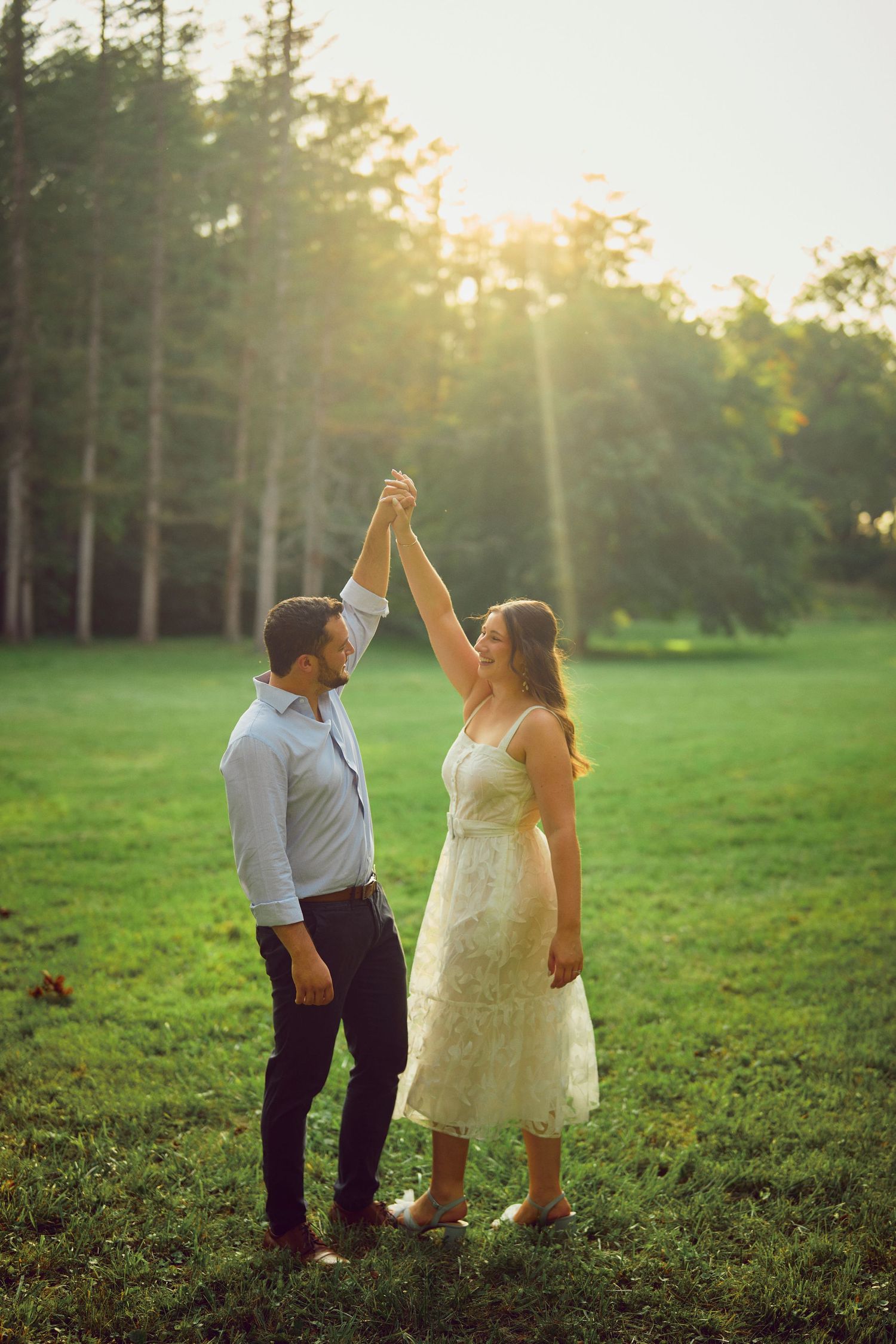 A couple dances together in a lush green field at sunset with sunbeams streaming through tall pine trees in the background.