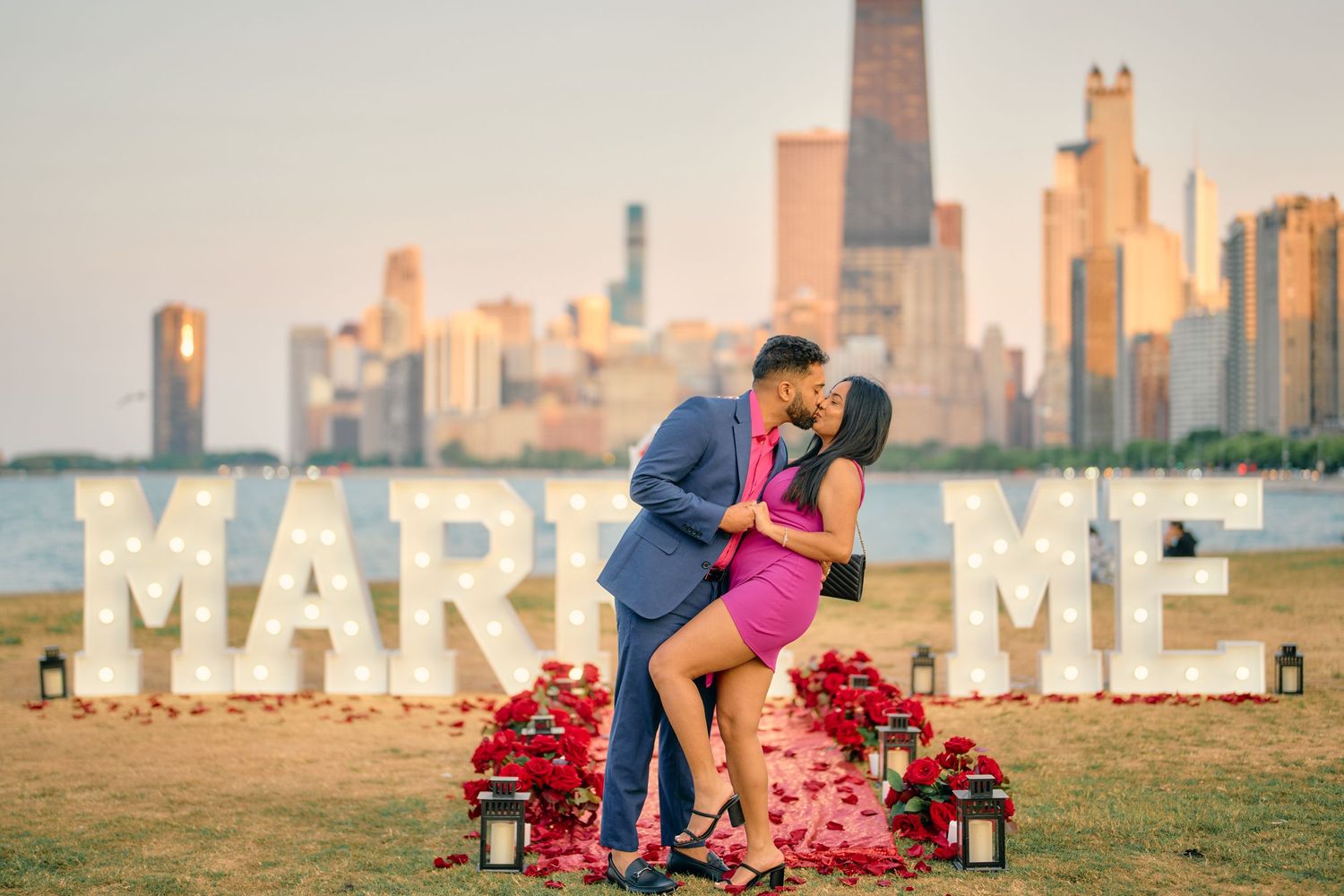 A couple shares a romantic moment in front of large 'MARRY ME' letters with Chicago skyline in the background at sunset.
