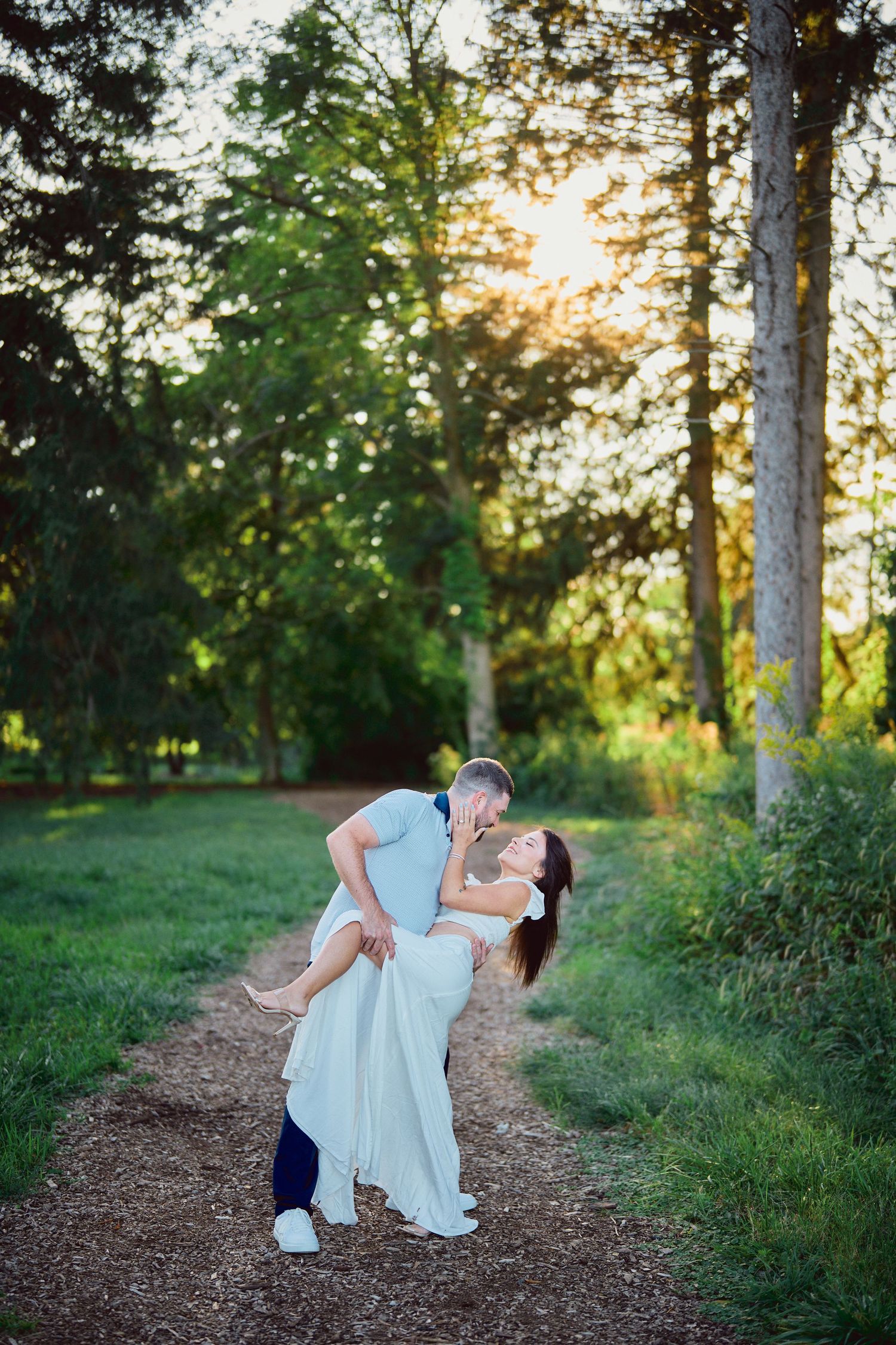 A romantic couple in light blue outfits share a tender moment on a wooded path at sunset.