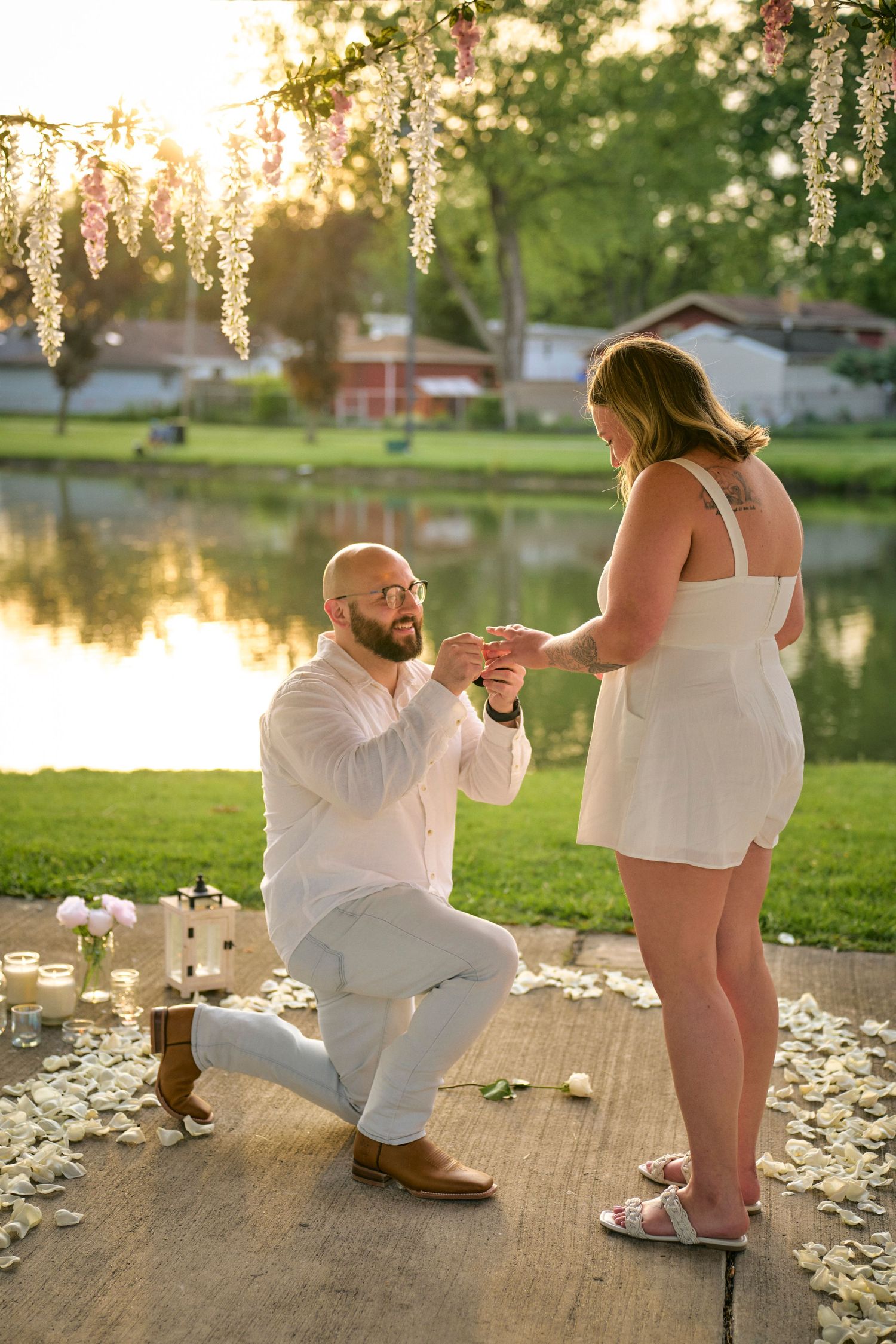 A romantic proposal scene at sunset by a lake with hanging lights and rose petals scattered on a wooden platform.