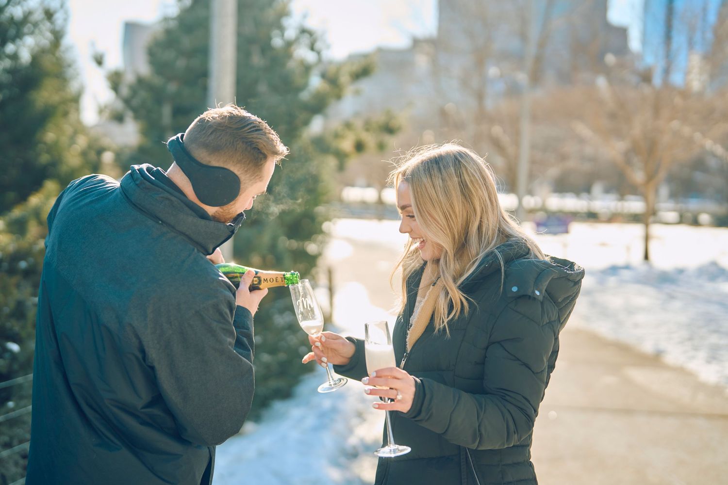 Two people share a warm moment together on a winter path as sunlight filters through trees in the background.