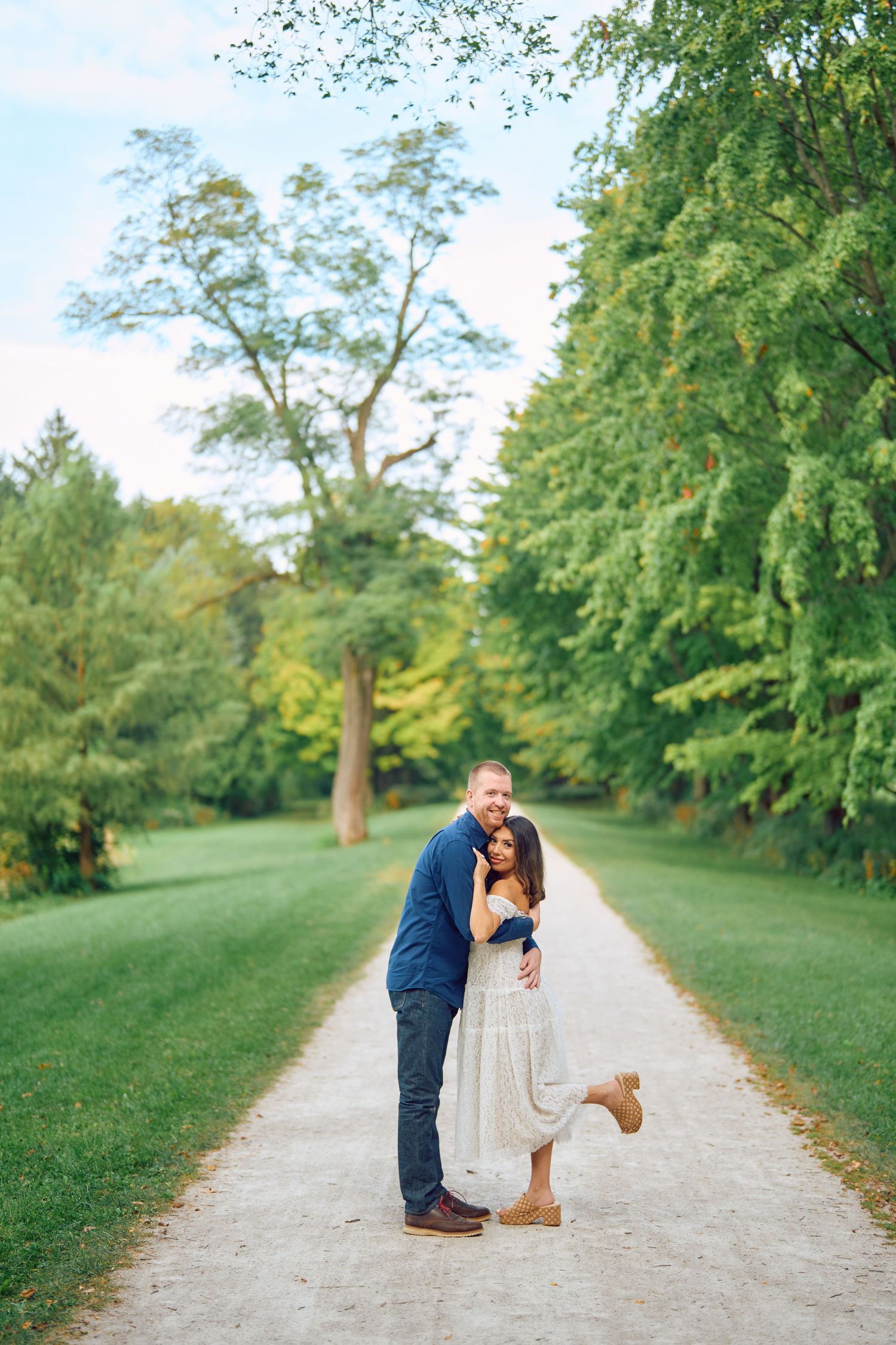 A romantic couple embraces on a tree-lined path in a lush green park setting on a sunny day.