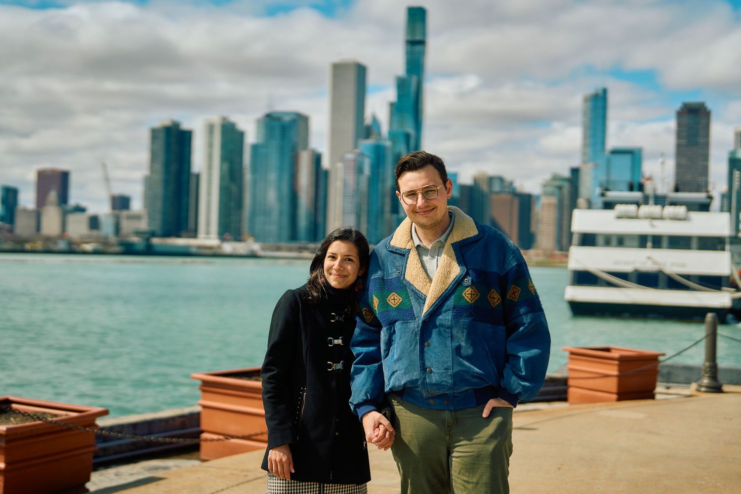 Two people in warm jackets stand at Navy Pier with the Chicago skyline and Lake Michigan visible in the background.