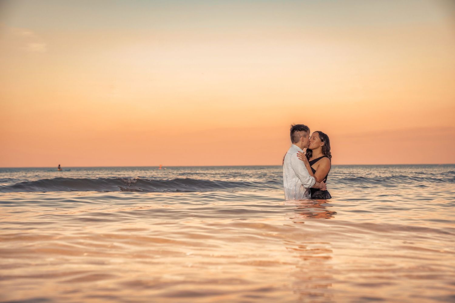 A romantic couple embraces in shallow ocean water during a golden sunset.