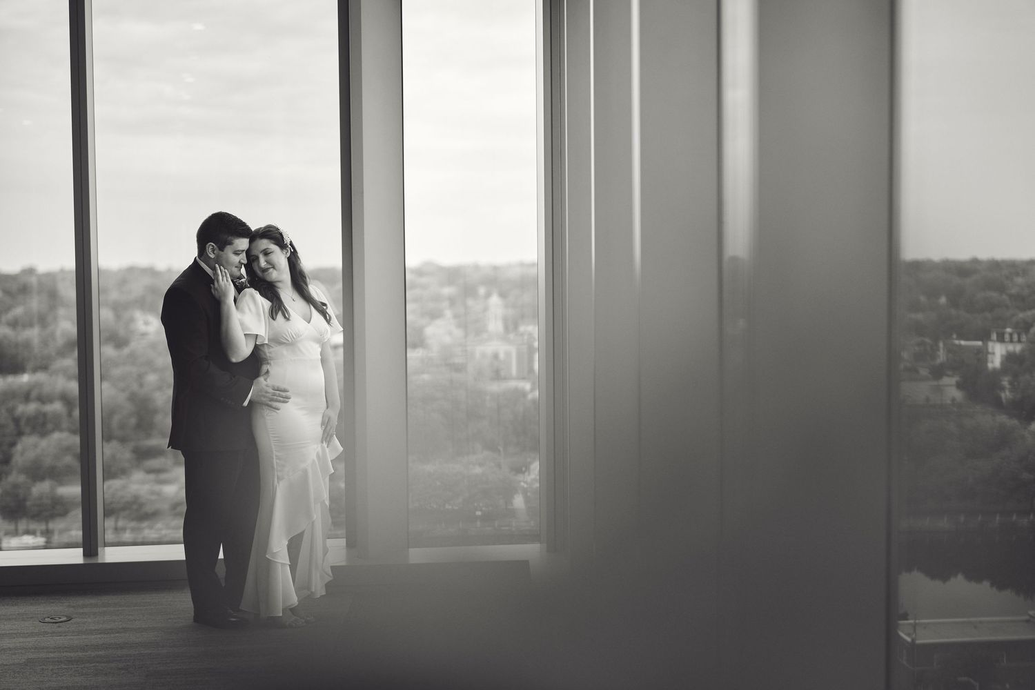 Black and white photograph of couple in formal attire standing beside window with scenic cityscape view.