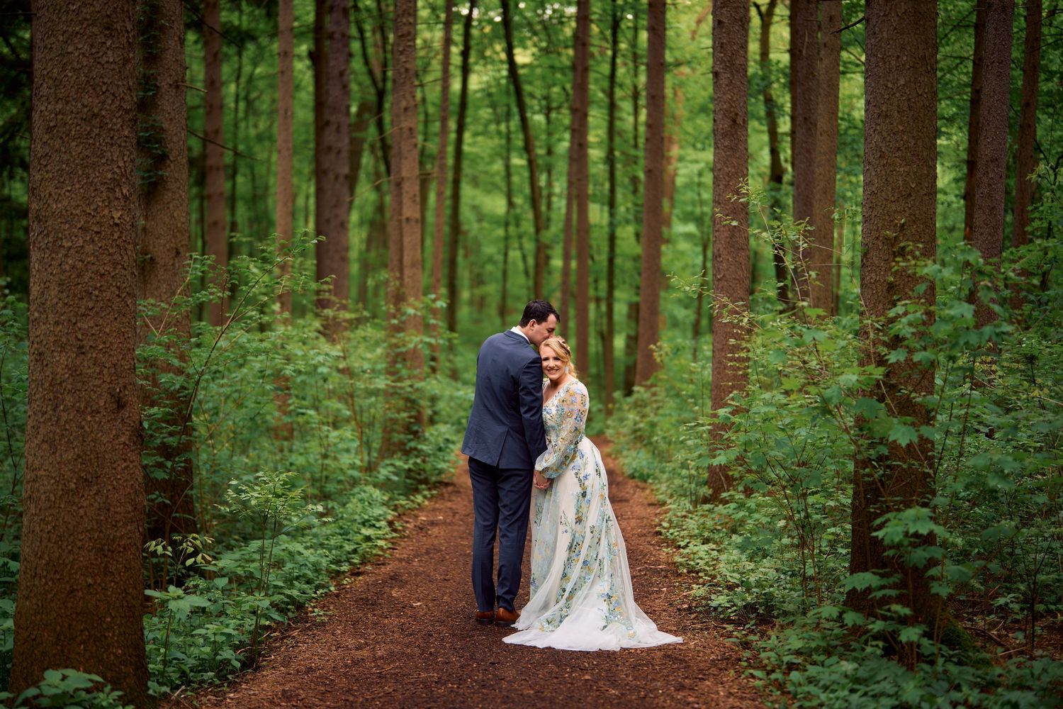 A couple shares a romantic moment on a dirt path through a lush green forest in elegant wedding attire.