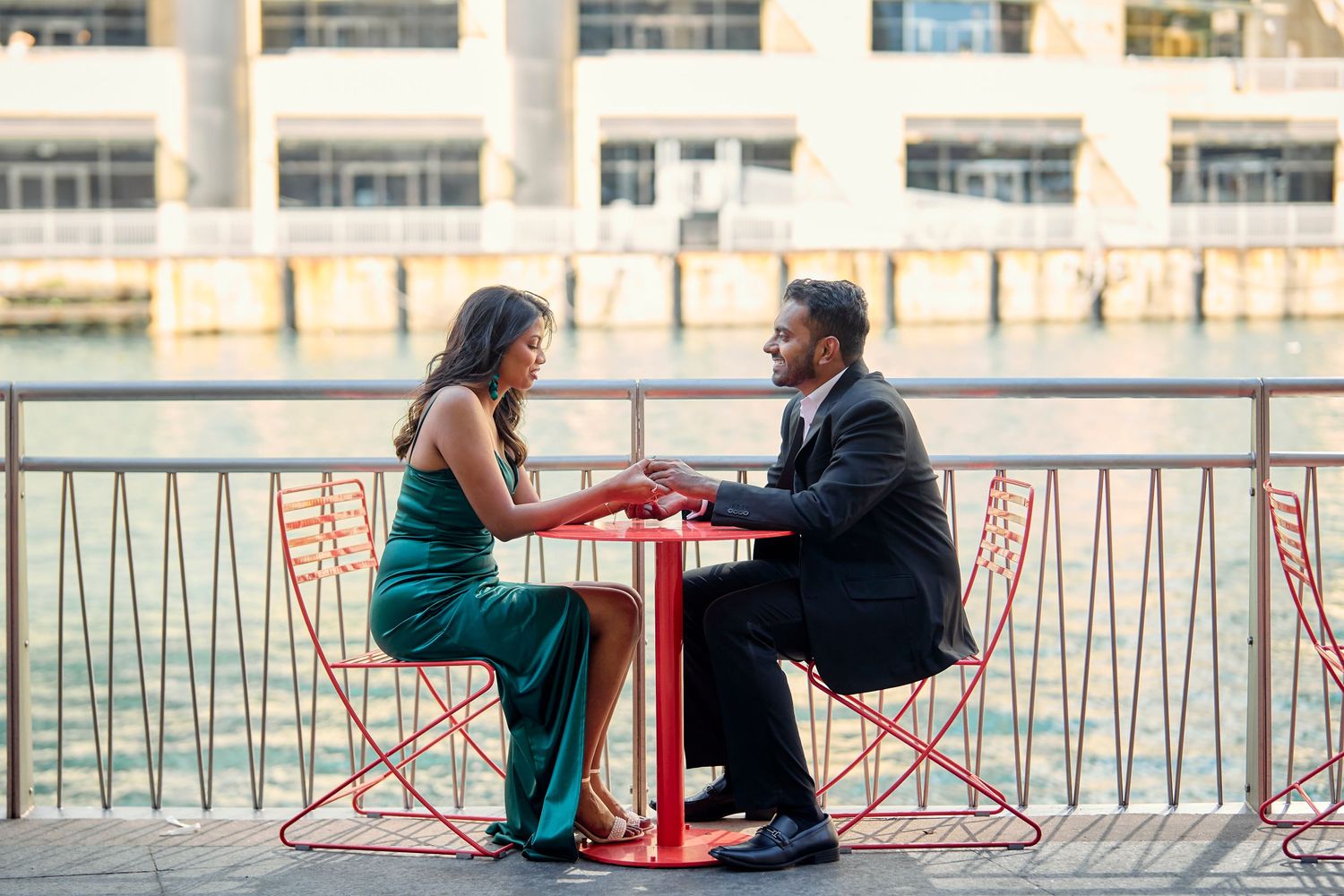 Couple sits at red metal table having romantic outdoor riverside dinner at dusk.