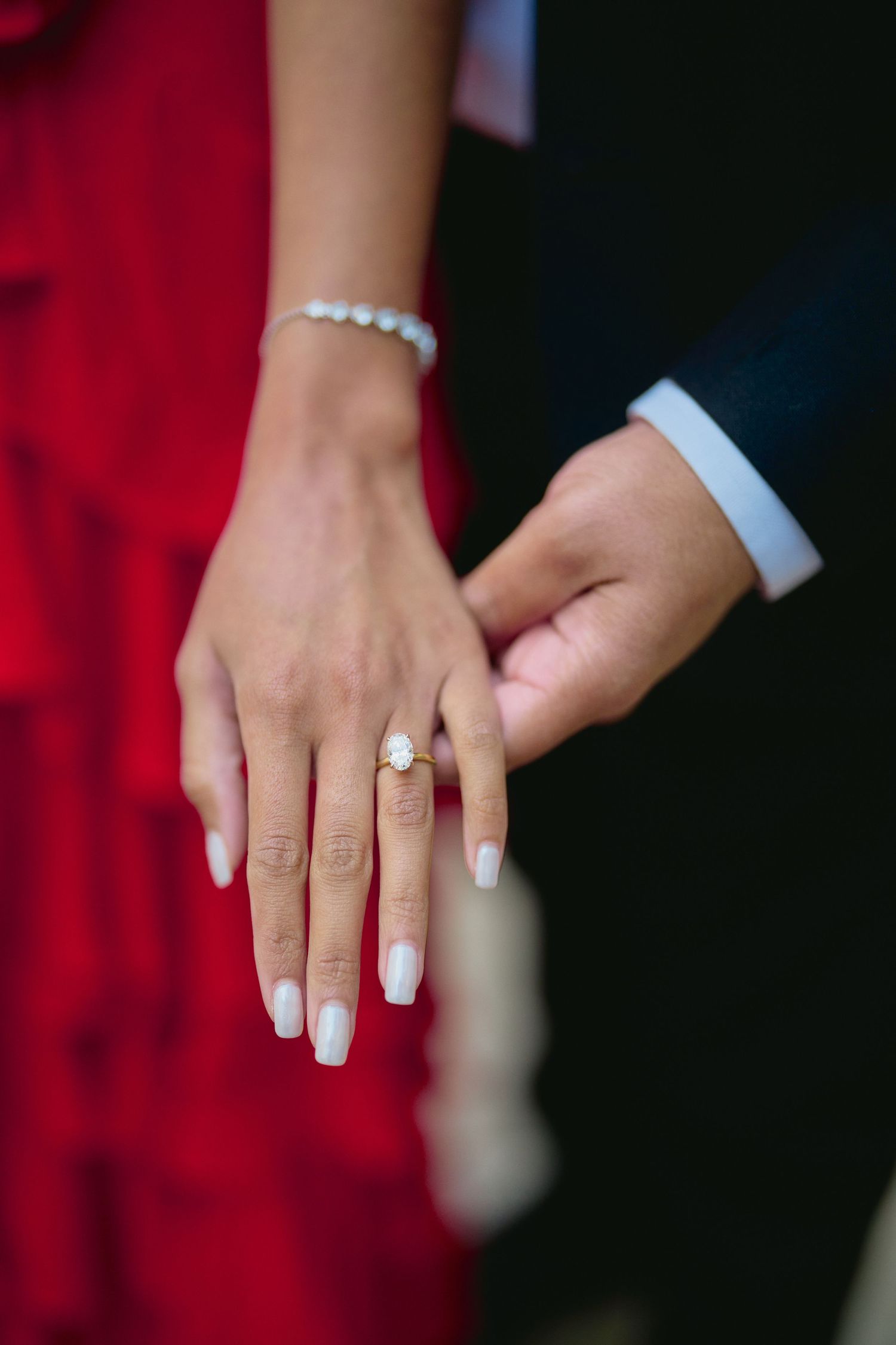 Close-up of hands showing a delicate engagement ring with white manicured nails and pearl bracelet details.