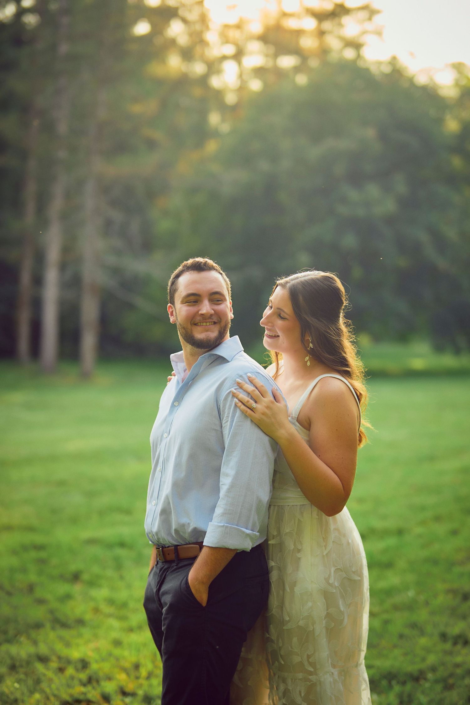 A couple shares a romantic moment together outdoors in a sunlit green field with trees in the background.
