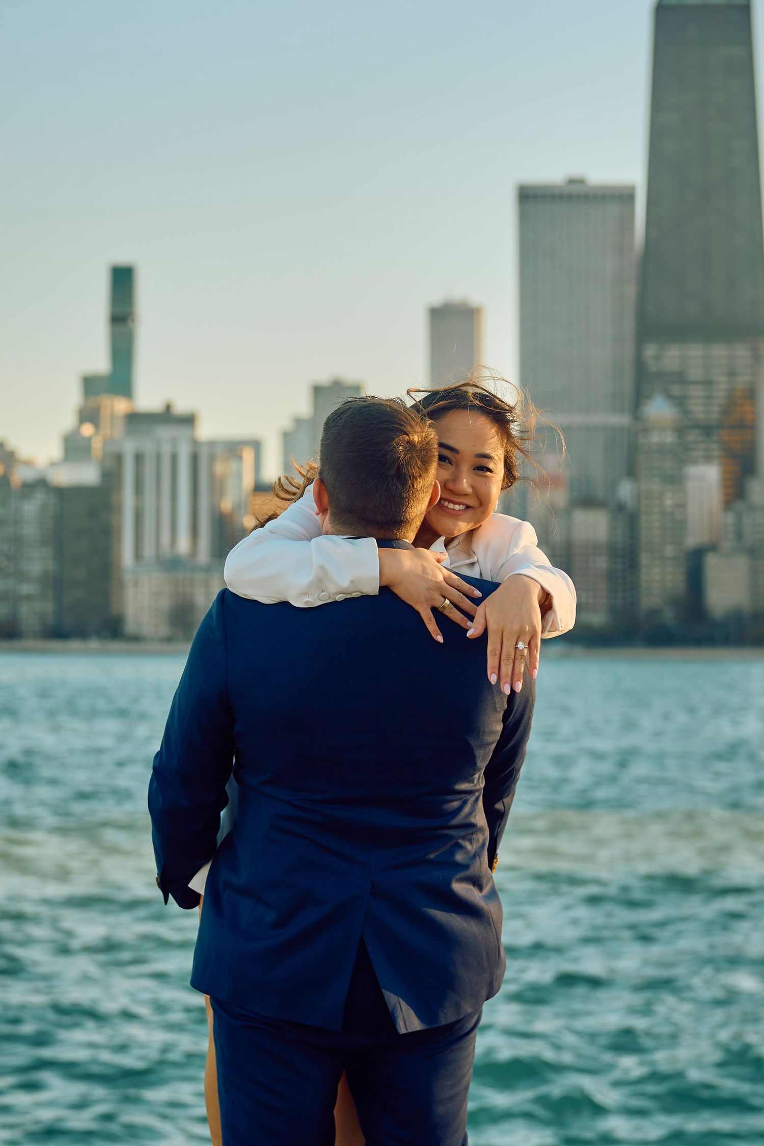 Two people embrace while standing by the water with a city skyline backdrop during golden hour in Chicago.