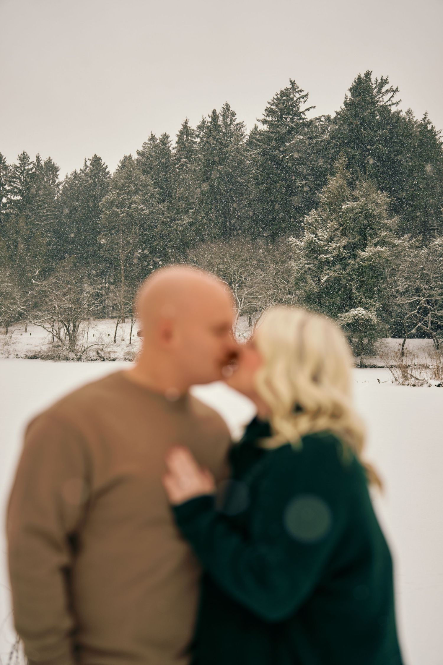 A couple shares a romantic winter kiss against a snowy forest background.