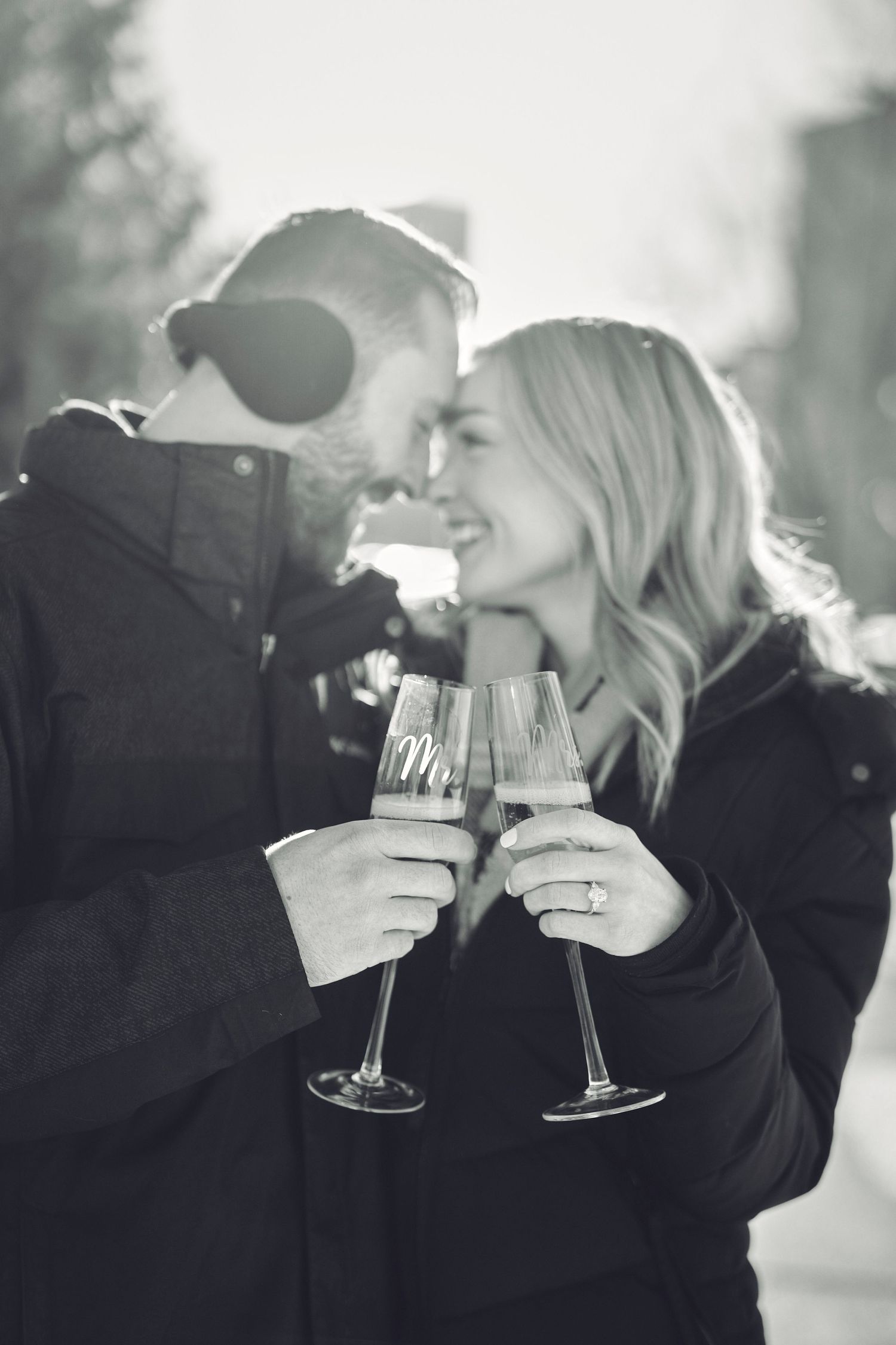 A couple in black winter coats share an intimate moment while toasting with wine glasses outdoors.