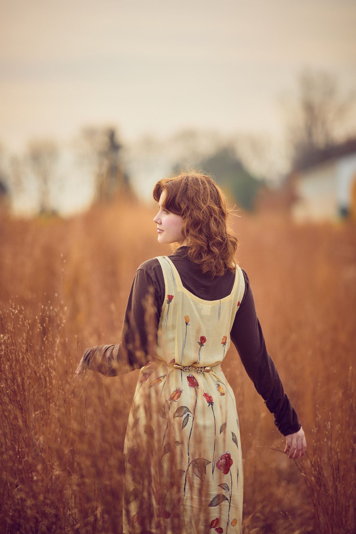 Person in floral dress stands in golden field at sunset, viewed from behind with long flowing hair.