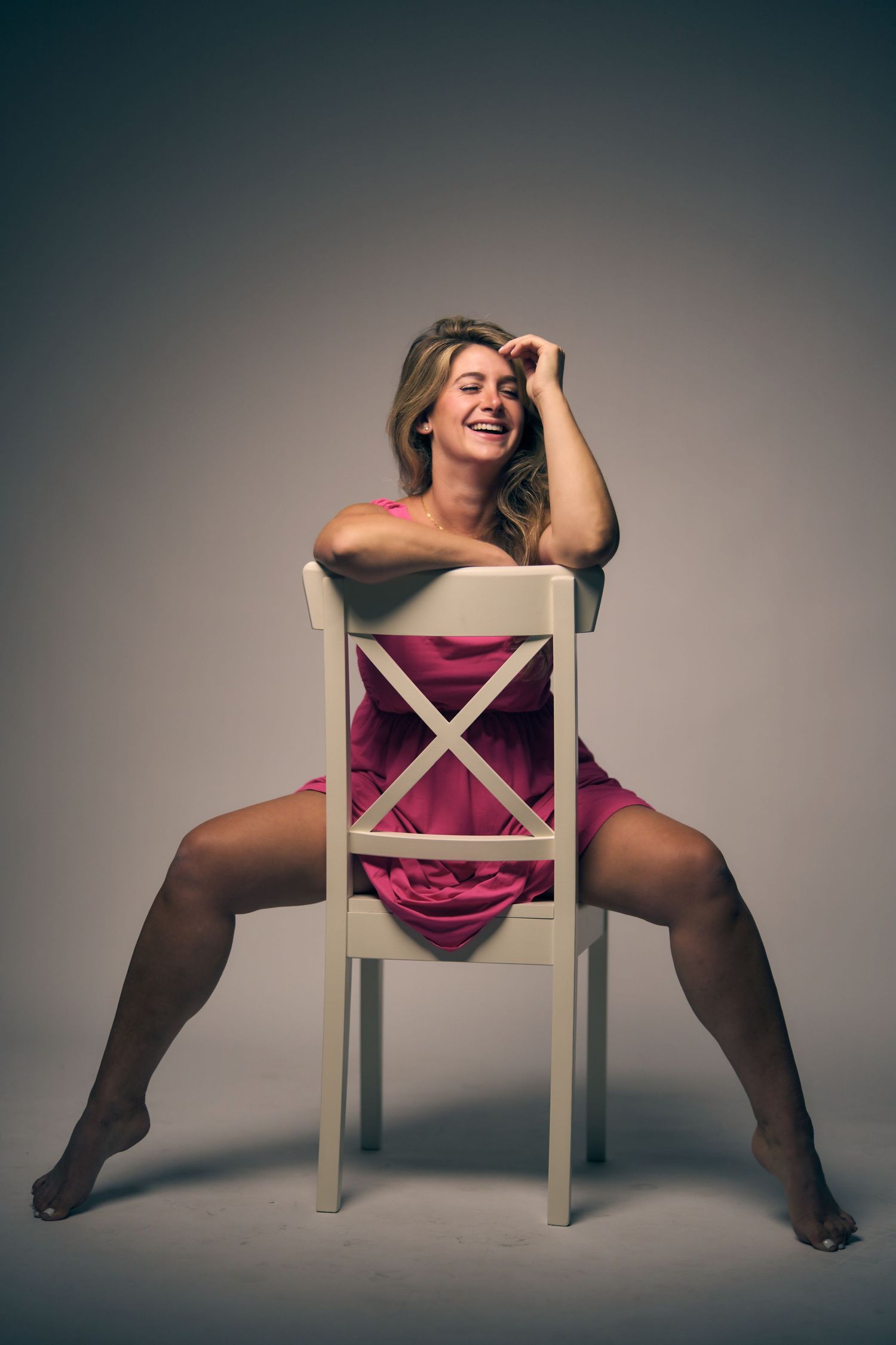 A figure poses playfully on a white chair against a dark backdrop in studio lighting.
