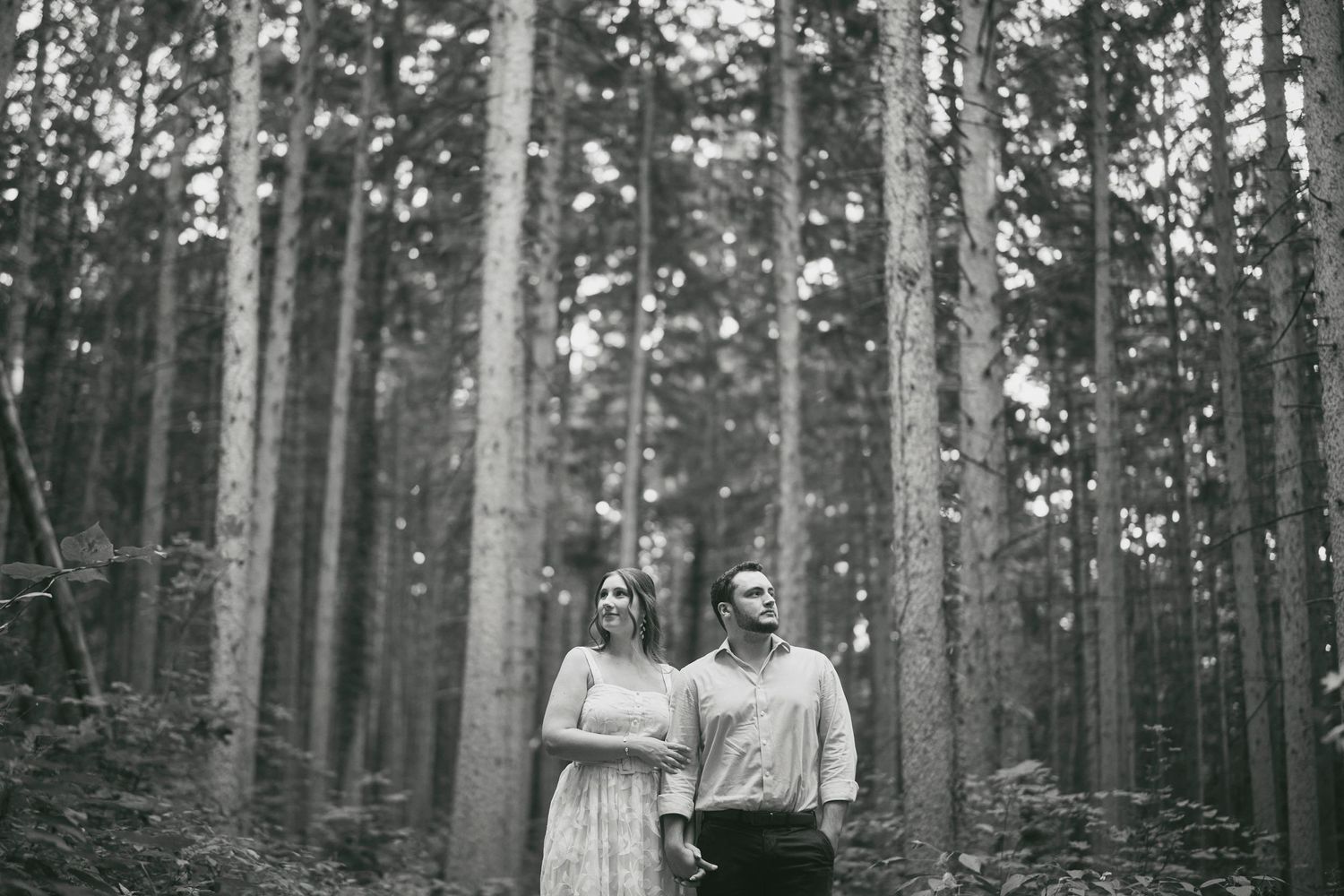 Black and white photo showing a couple standing together in a tranquil forest of tall pine trees.