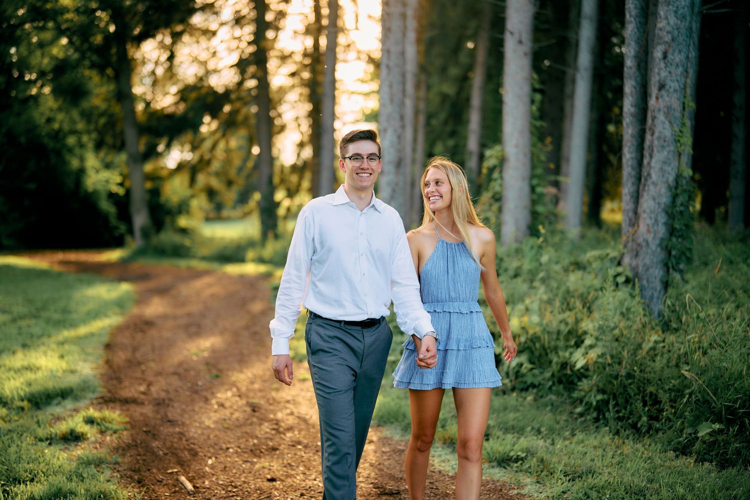A couple walks hand in hand along a sunlit forest path on a summer evening.