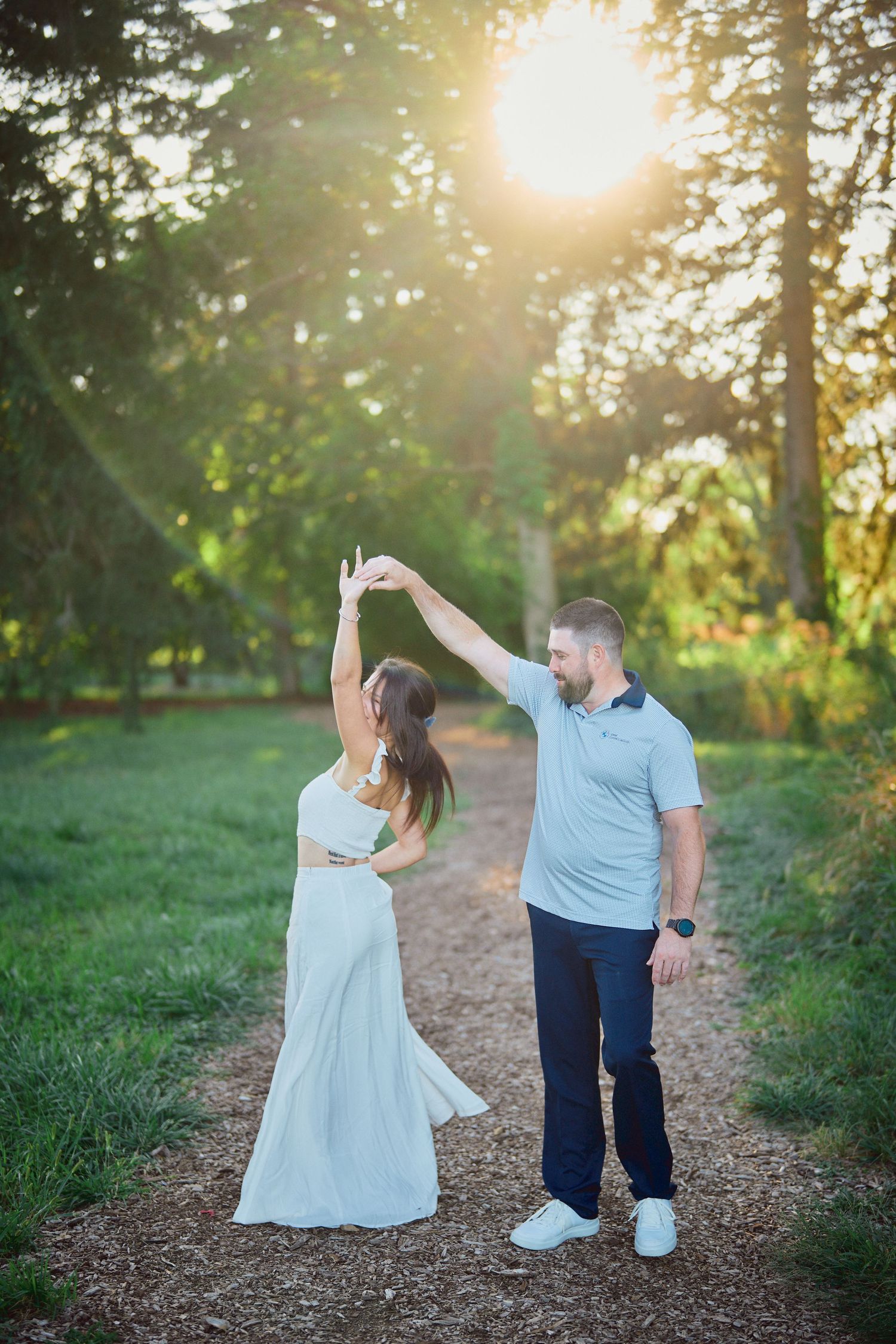Couple dancing together on a forest trail during golden hour sunset while wearing blue and white outfits.