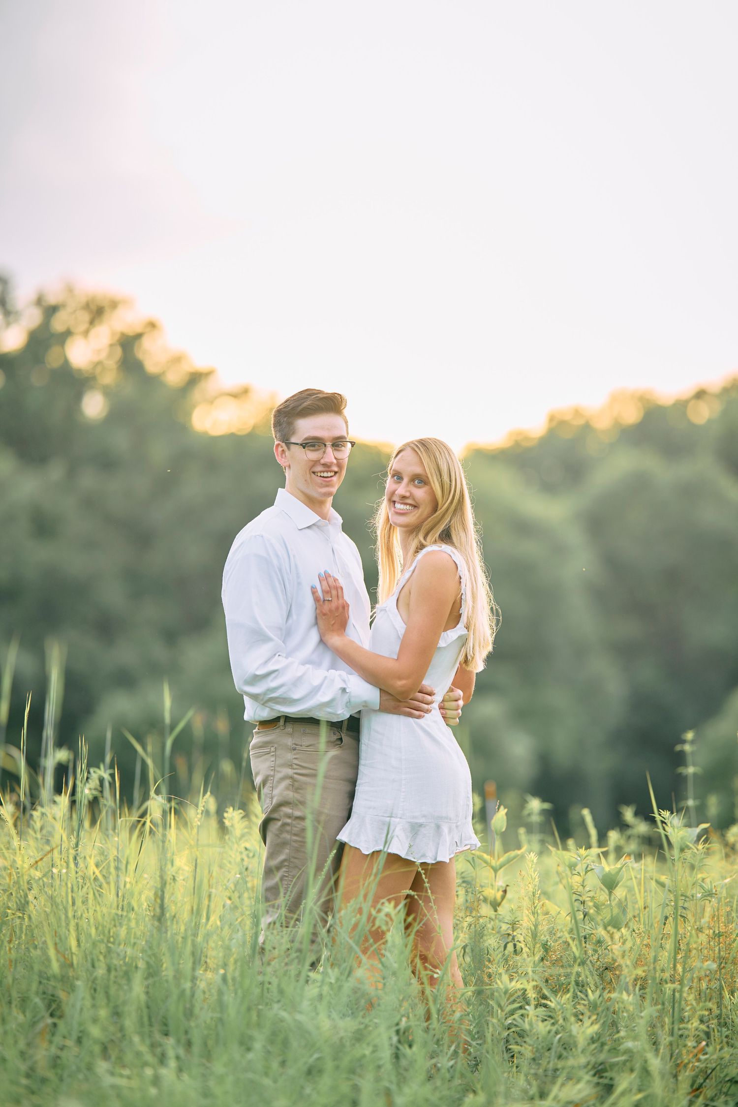 A romantic couple embraces in a sunlit meadow during golden hour, wearing white and khaki casual attire.