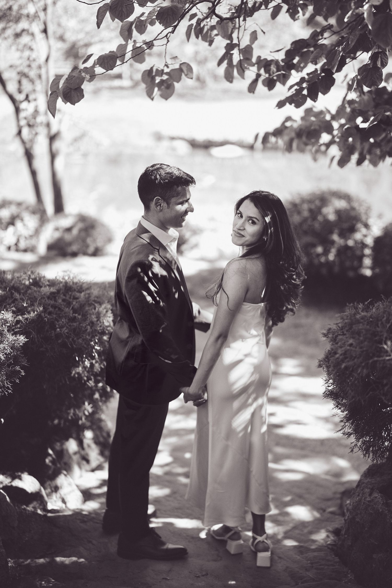 A black and white romantic photo of a couple standing together on a garden path under dappled sunlight.
