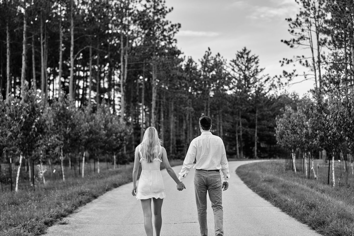 Black and white photo of a couple walking hand in hand down a tree-lined rural path.