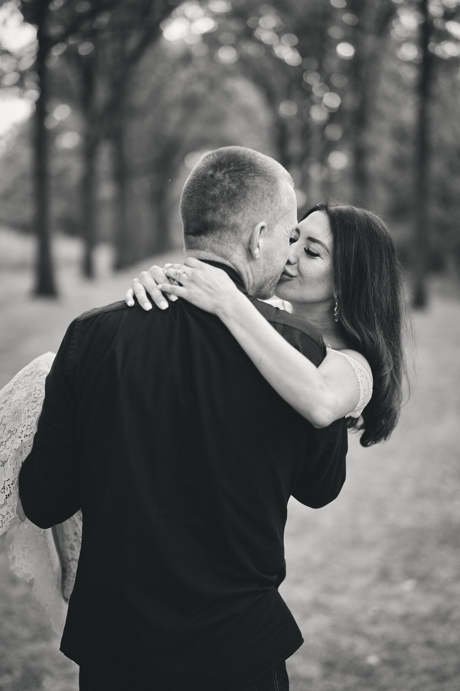 A tender black and white moment of a couple sharing an intimate embrace in a tree-lined park setting.