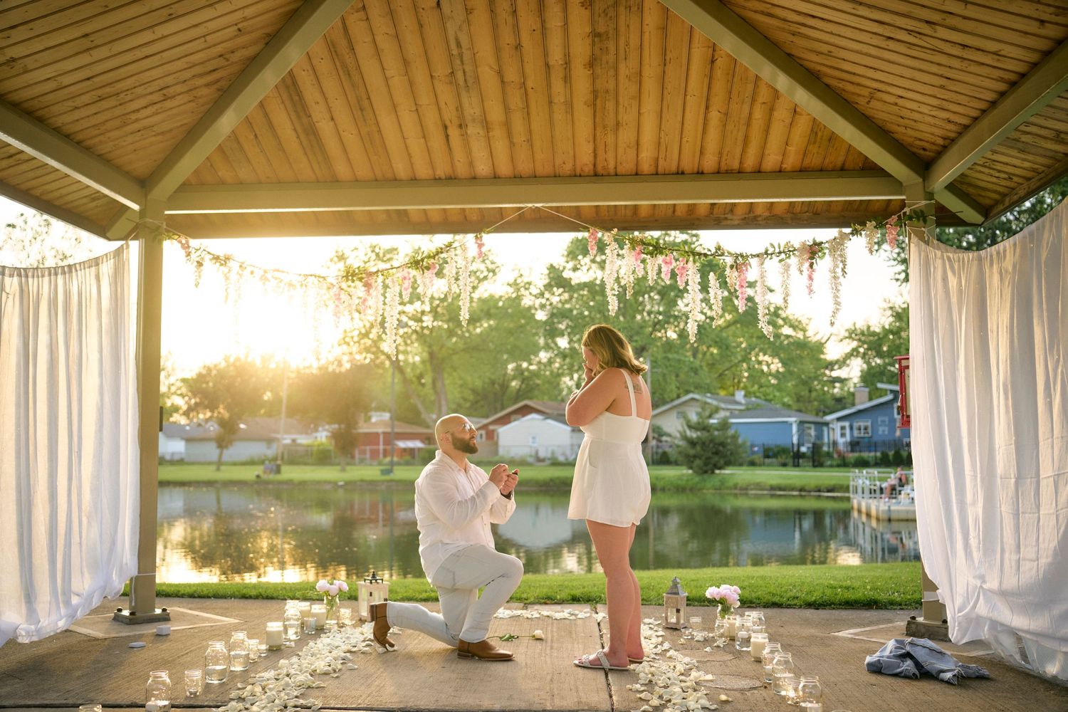 A romantic proposal at sunset under a waterfront gazebo decorated with white curtains and flower petals.