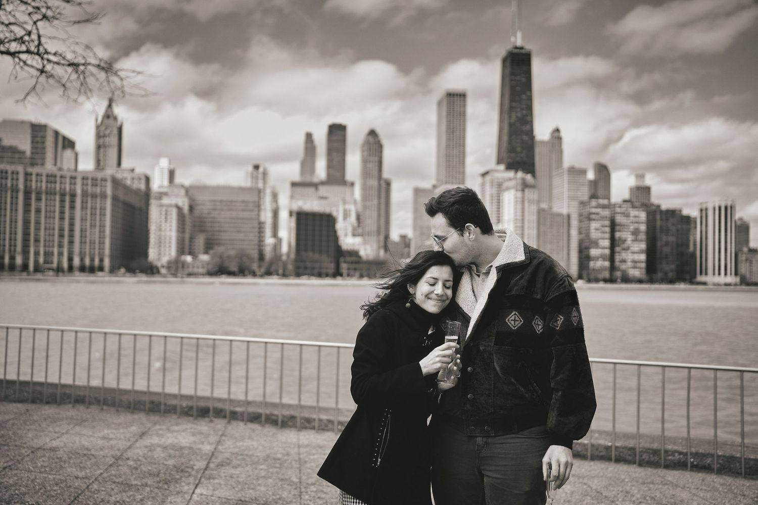 A romantic black and white scene of a couple embracing against the Chicago skyline on a cloudy day.
