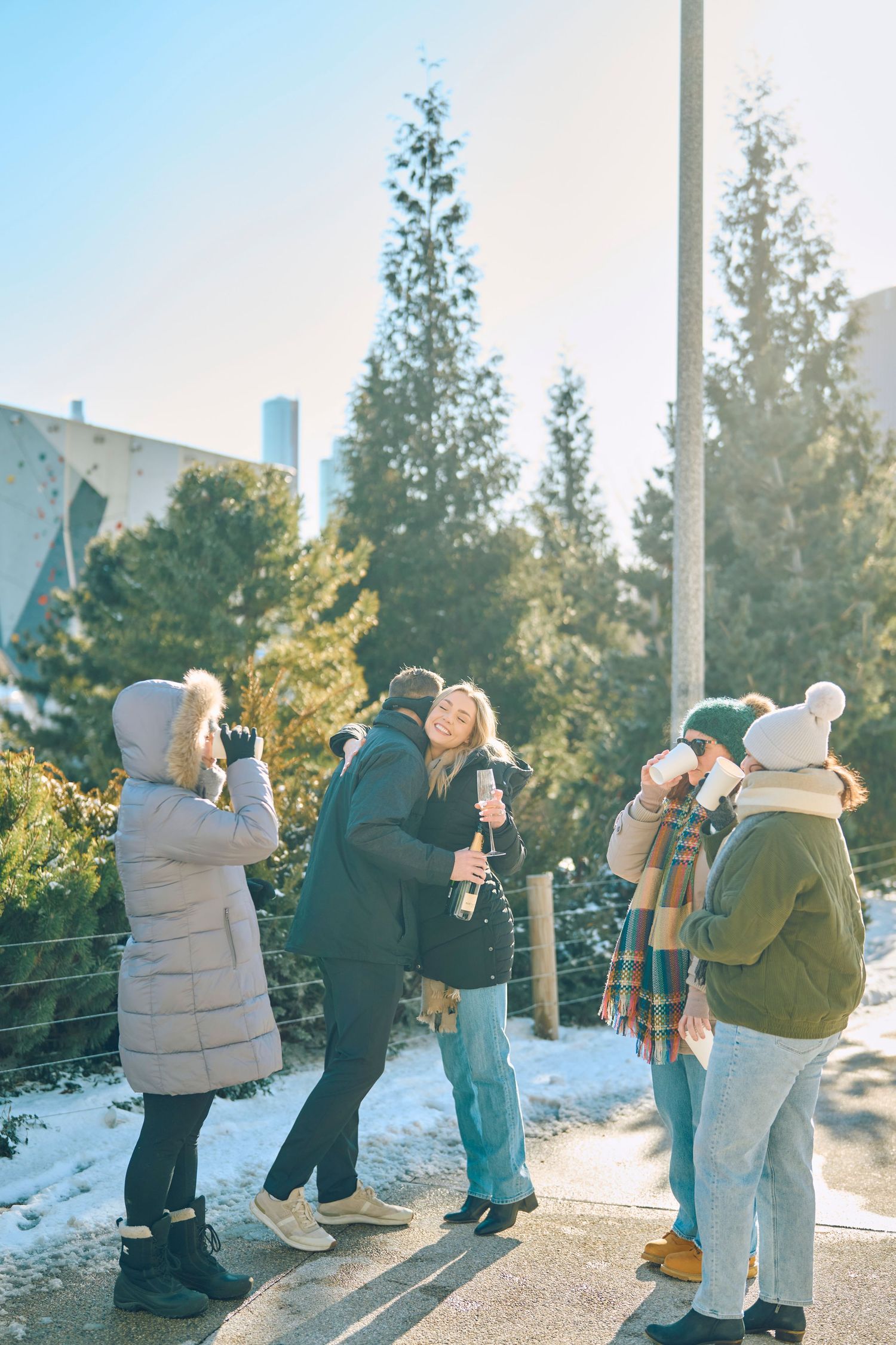 Friends bundled in winter coats take photos on a snowy path with evergreen trees in the background.