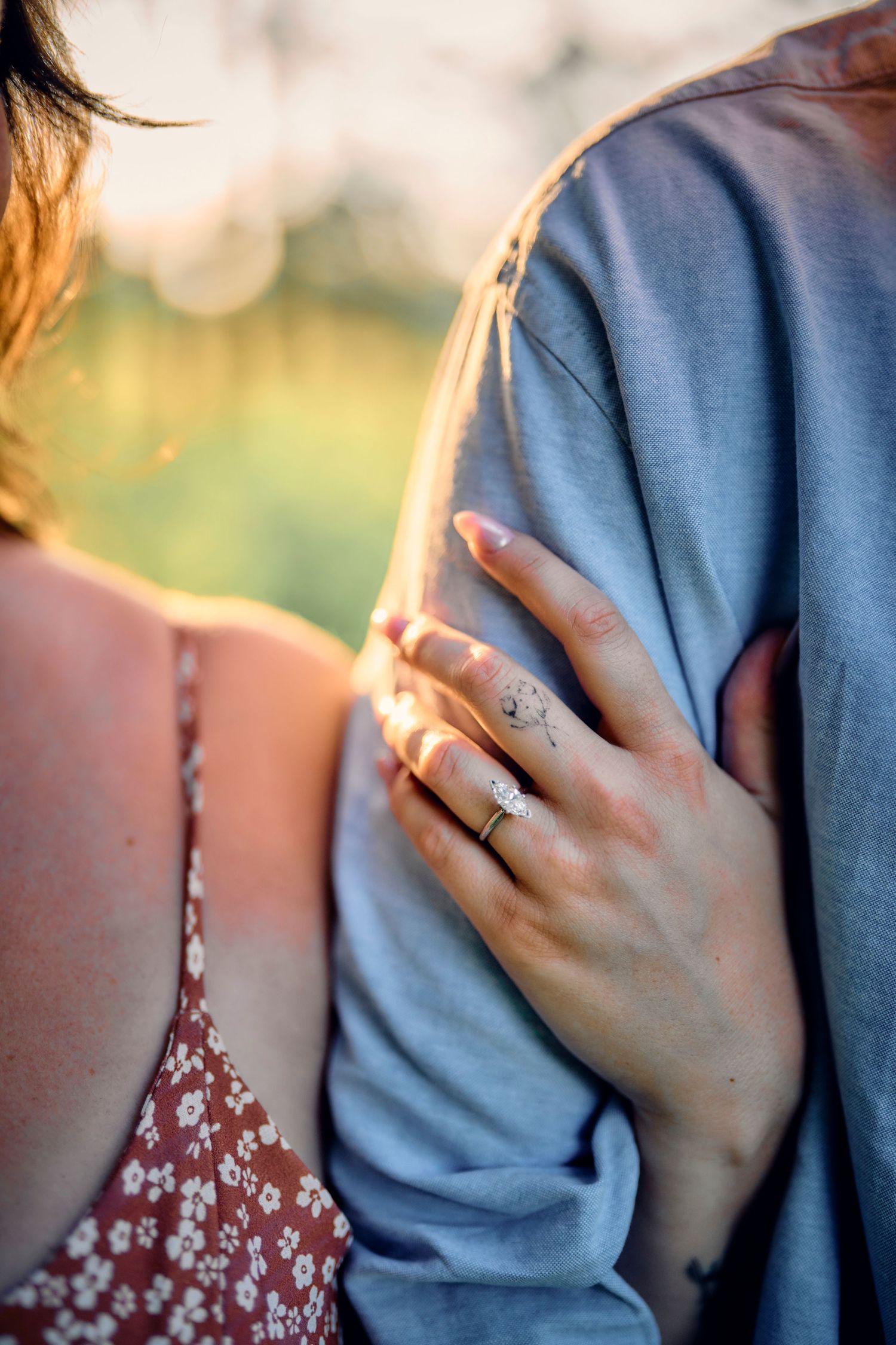 Close-up shot of a person's hand wearing an engagement ring resting on their partner's blue shirt at sunset.