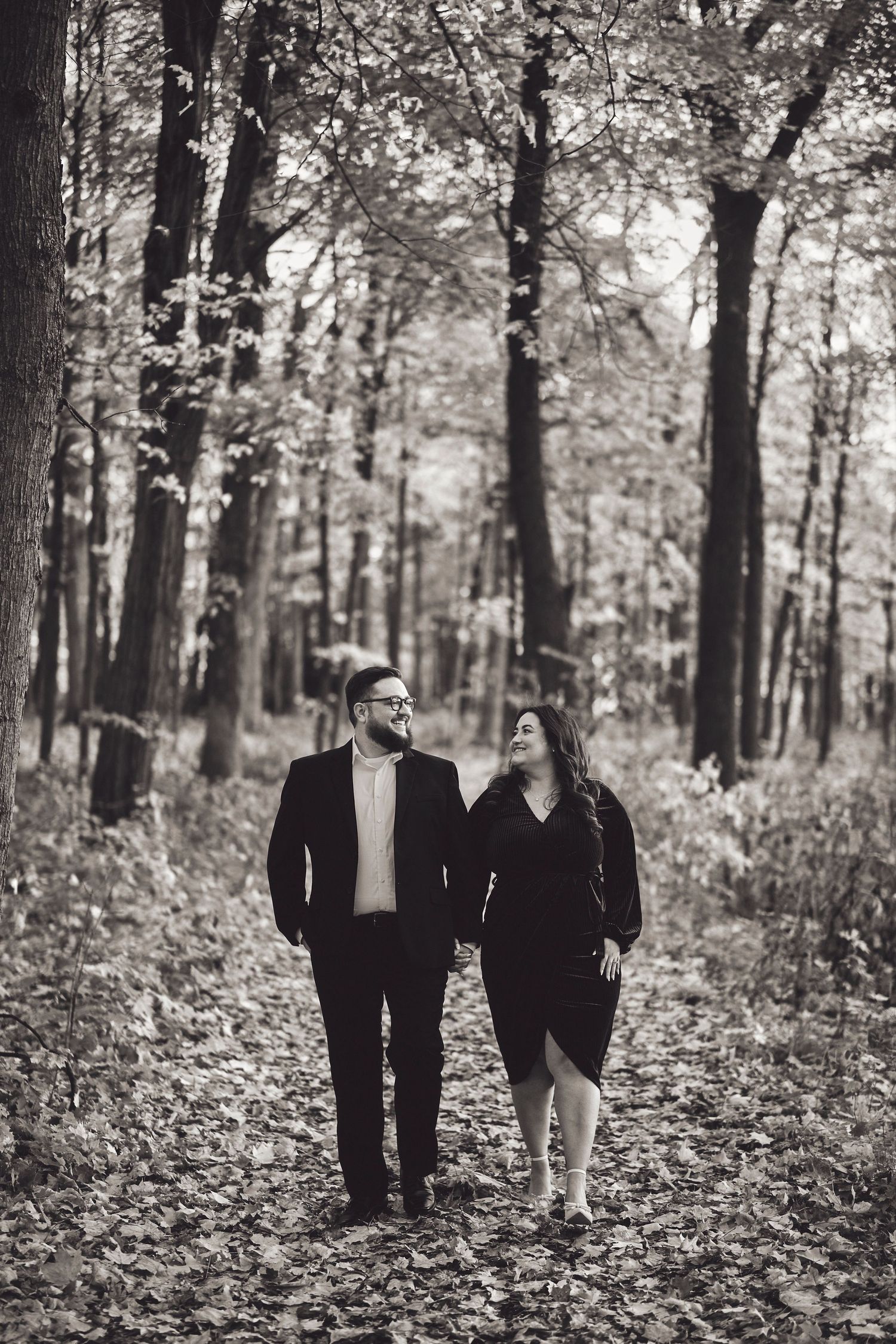 A couple walking together on a leaf-covered path through a serene autumn forest in black and white.