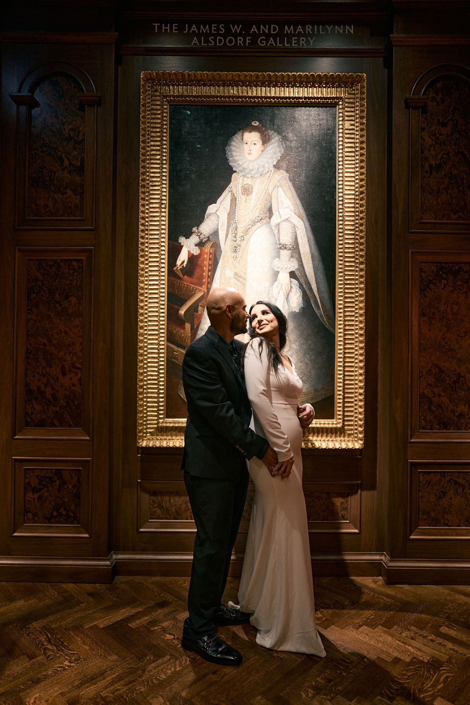 A couple in formal attire poses together in a museum gallery in front of a large ornate gold-framed classical portrait.