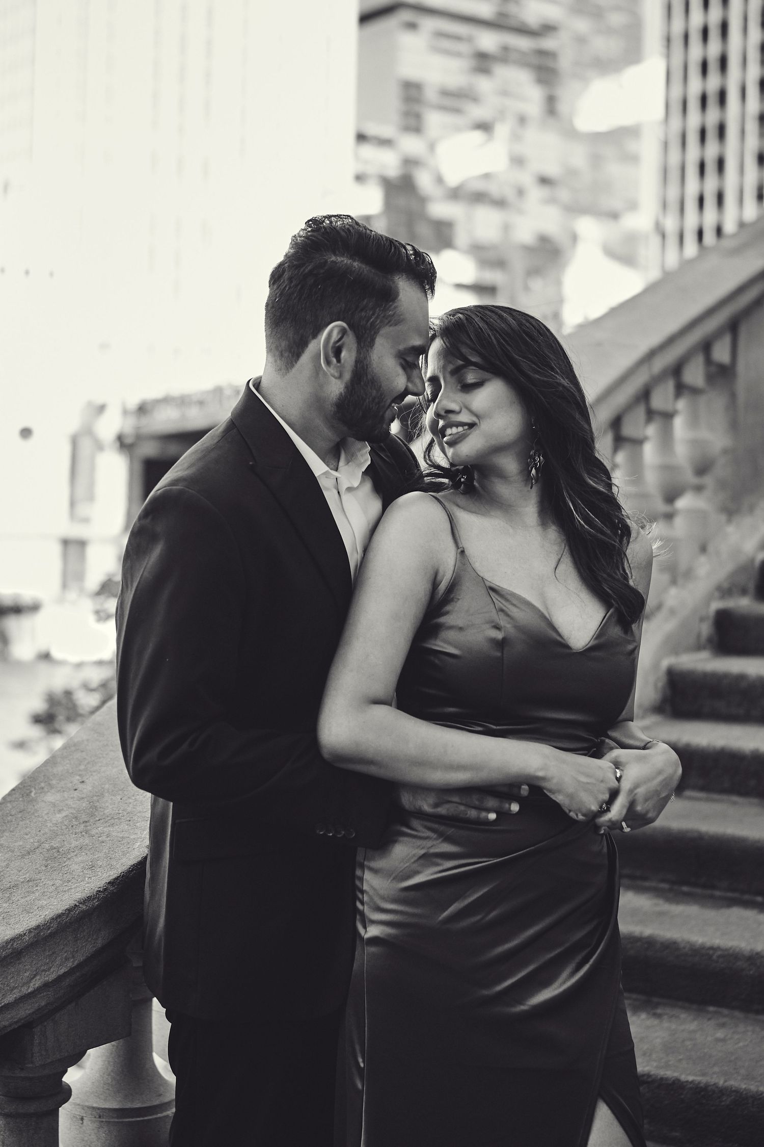 Elegant couple in formal attire share intimate black and white moment on urban steps.