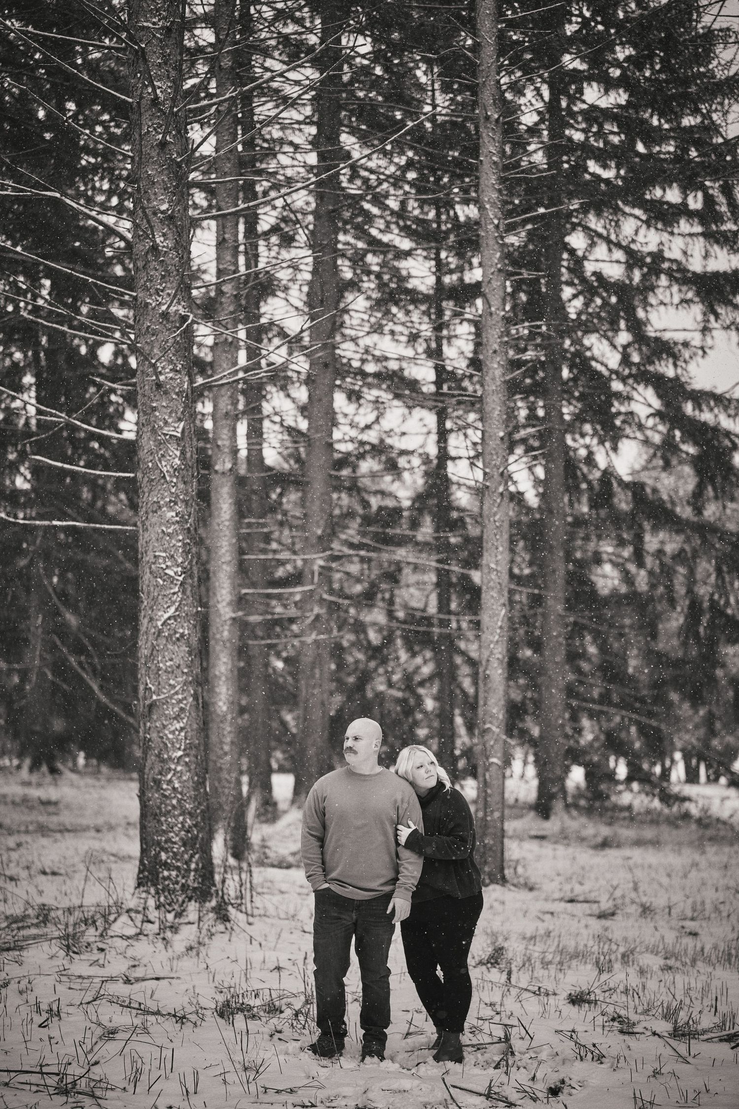 A couple walks together through a snowy forest of tall evergreen trees in black and white.
