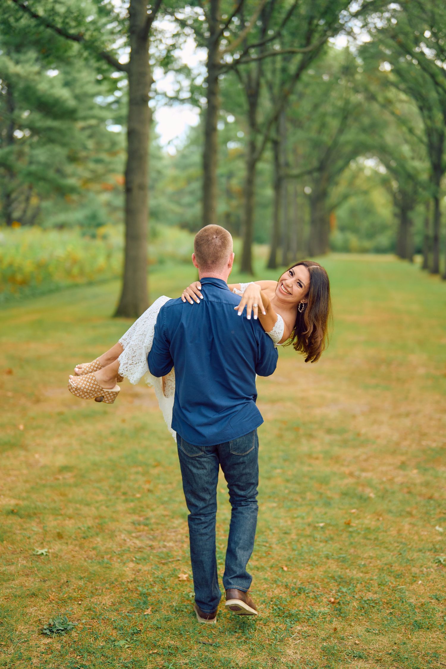 A couple shares a romantic moment on a tree-lined path as he carries her through a lush green park setting.