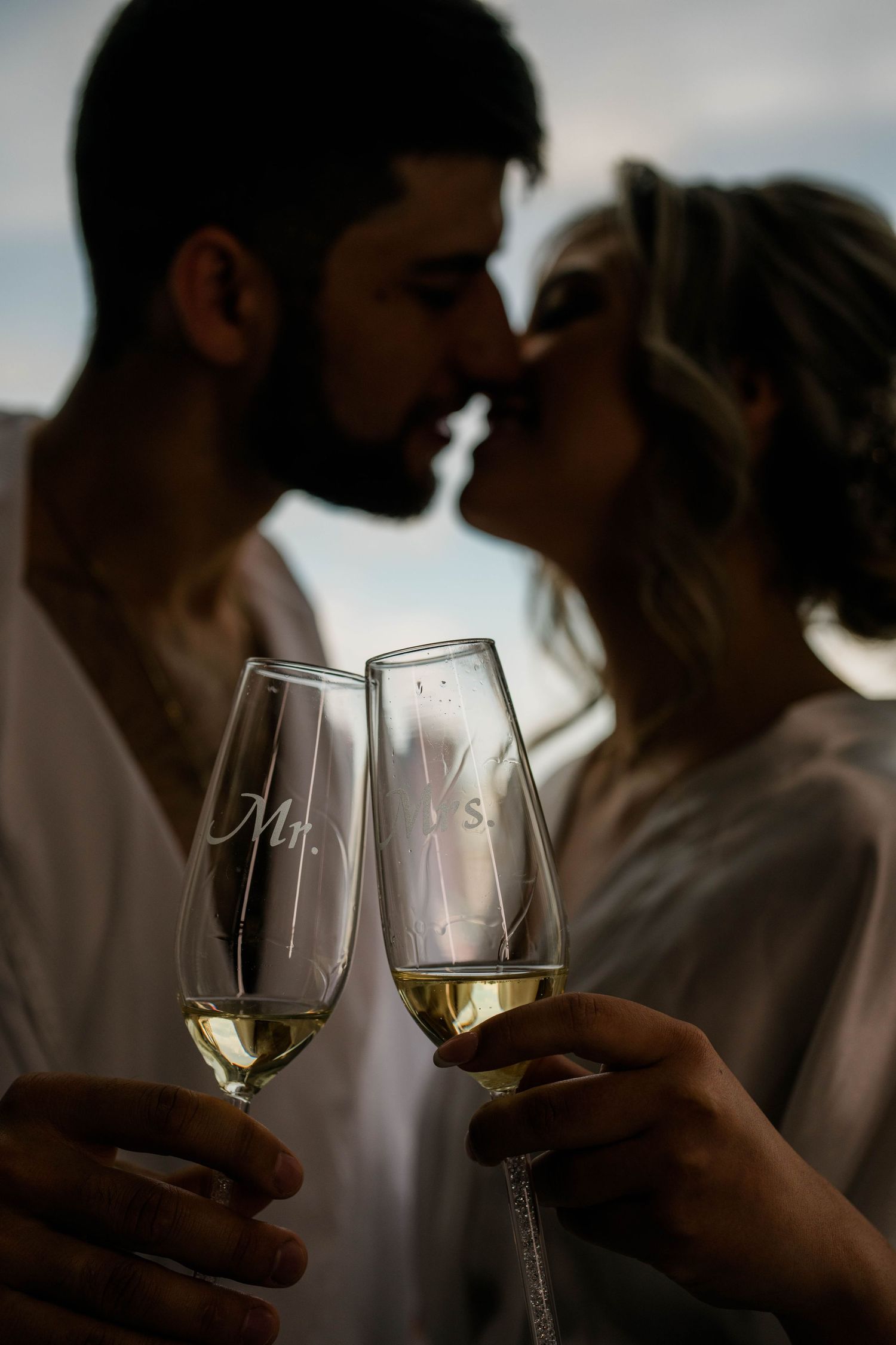 Two wine glasses filled with white wine clink together in front of a romantic silhouetted couple sharing a kiss.