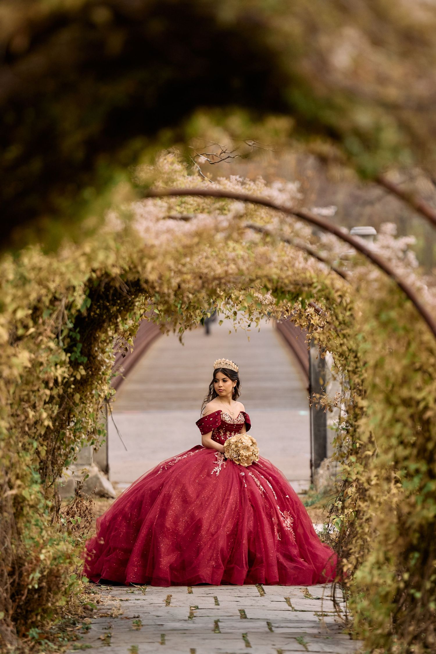 A stunning burgundy ballgown dress photographed through a garden archway covered in delicate vines and flowers.