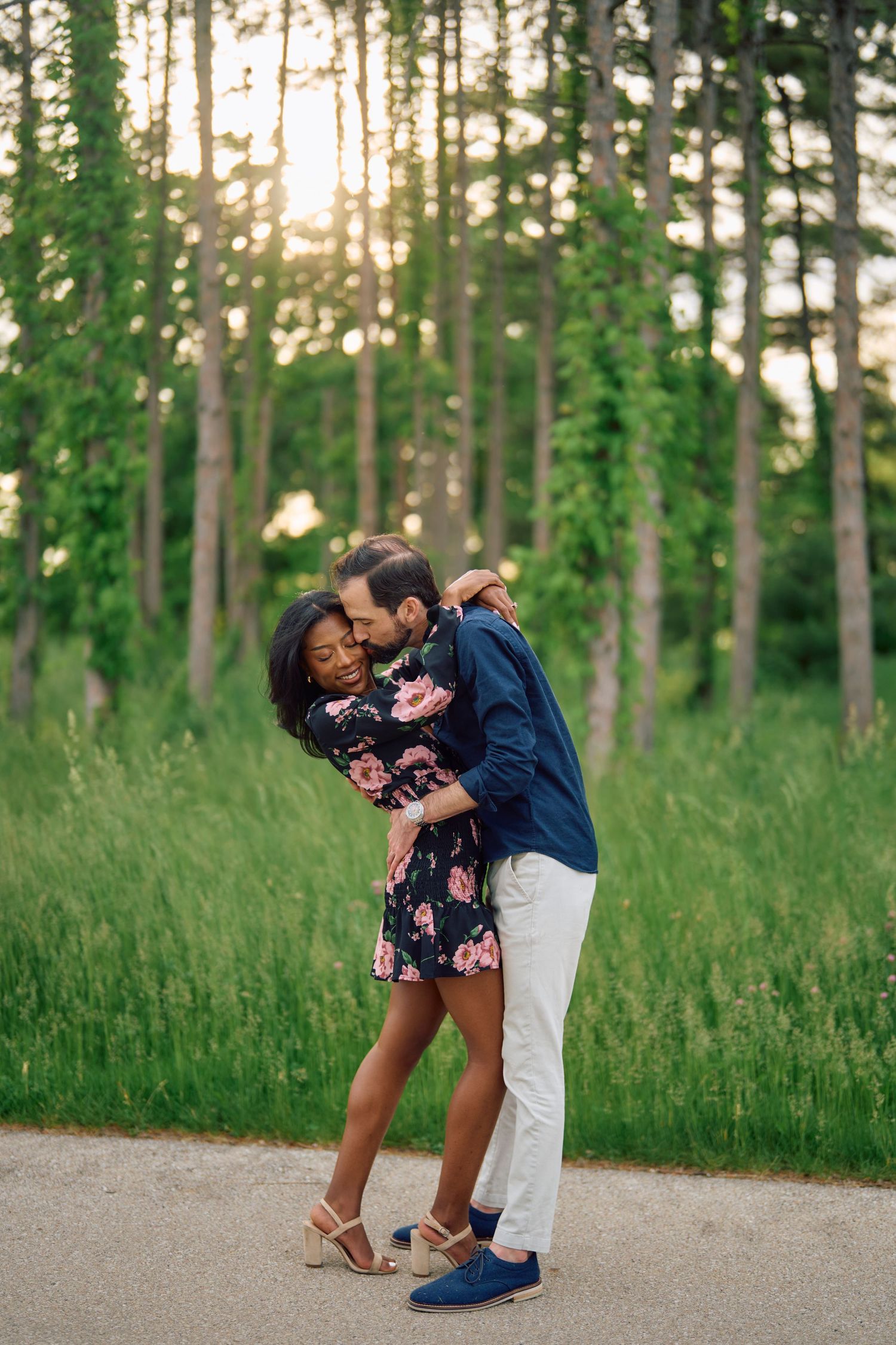 A couple shares an intimate moment on a path surrounded by tall trees and golden sunlight.