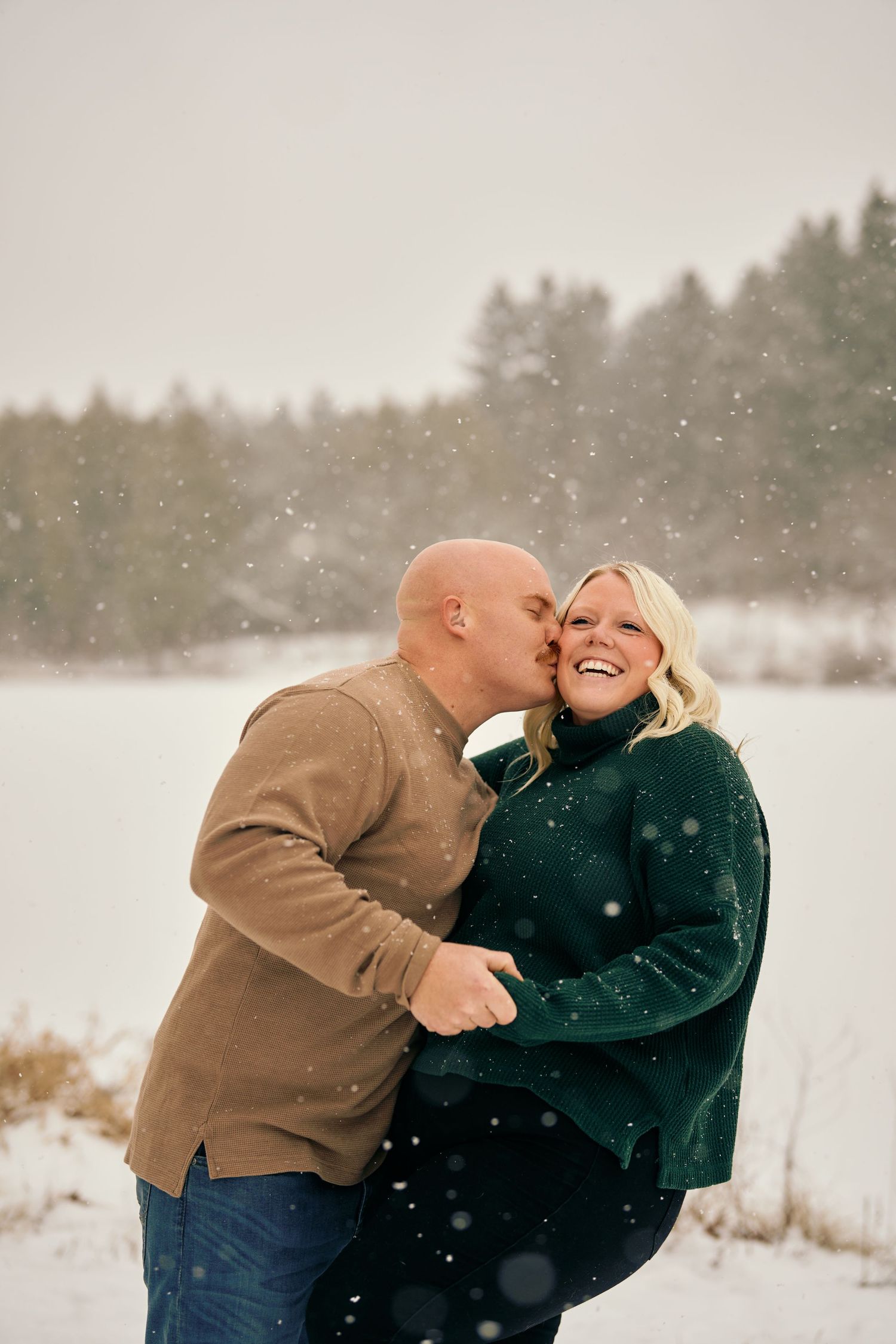 Couple shares a tender moment laughing together in the falling snow on a winter day by a frozen lake.