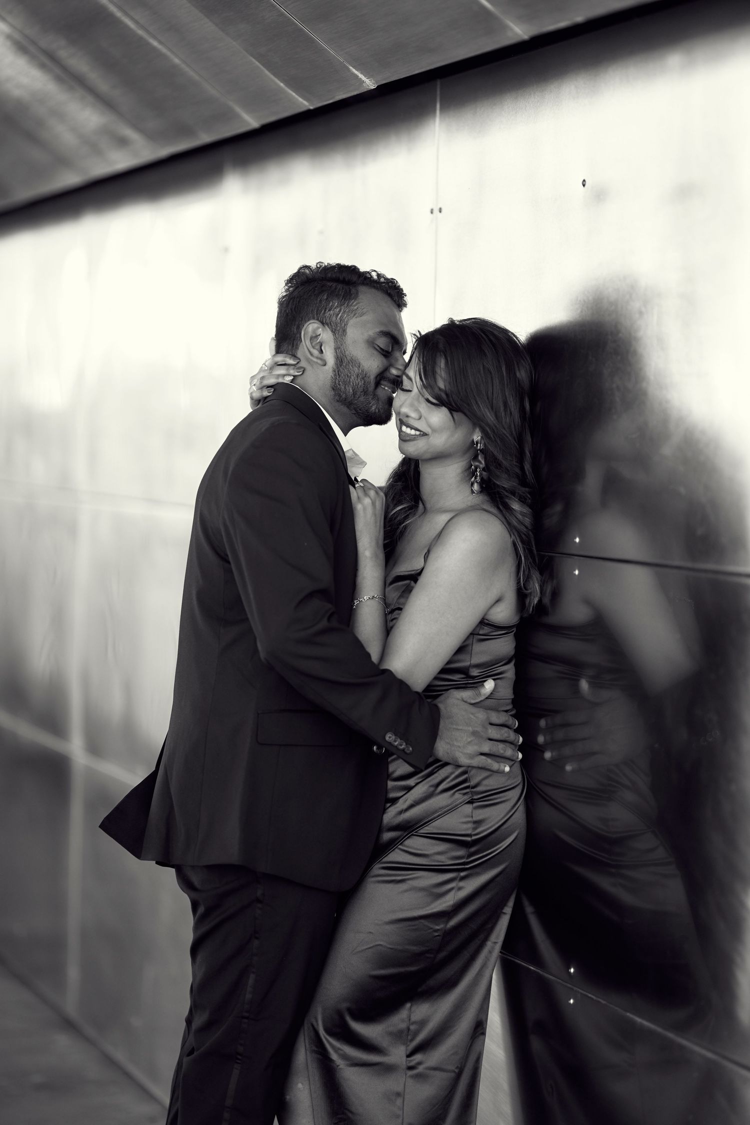 A romantic black and white portrait of a couple in formal attire embracing against a shadowy wall.