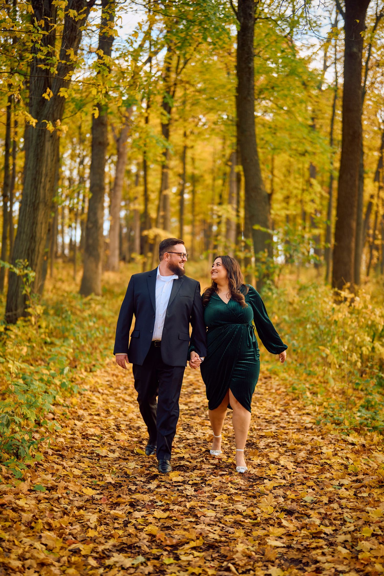 A couple walks hand in hand along a leaf-covered path through a golden autumn forest.