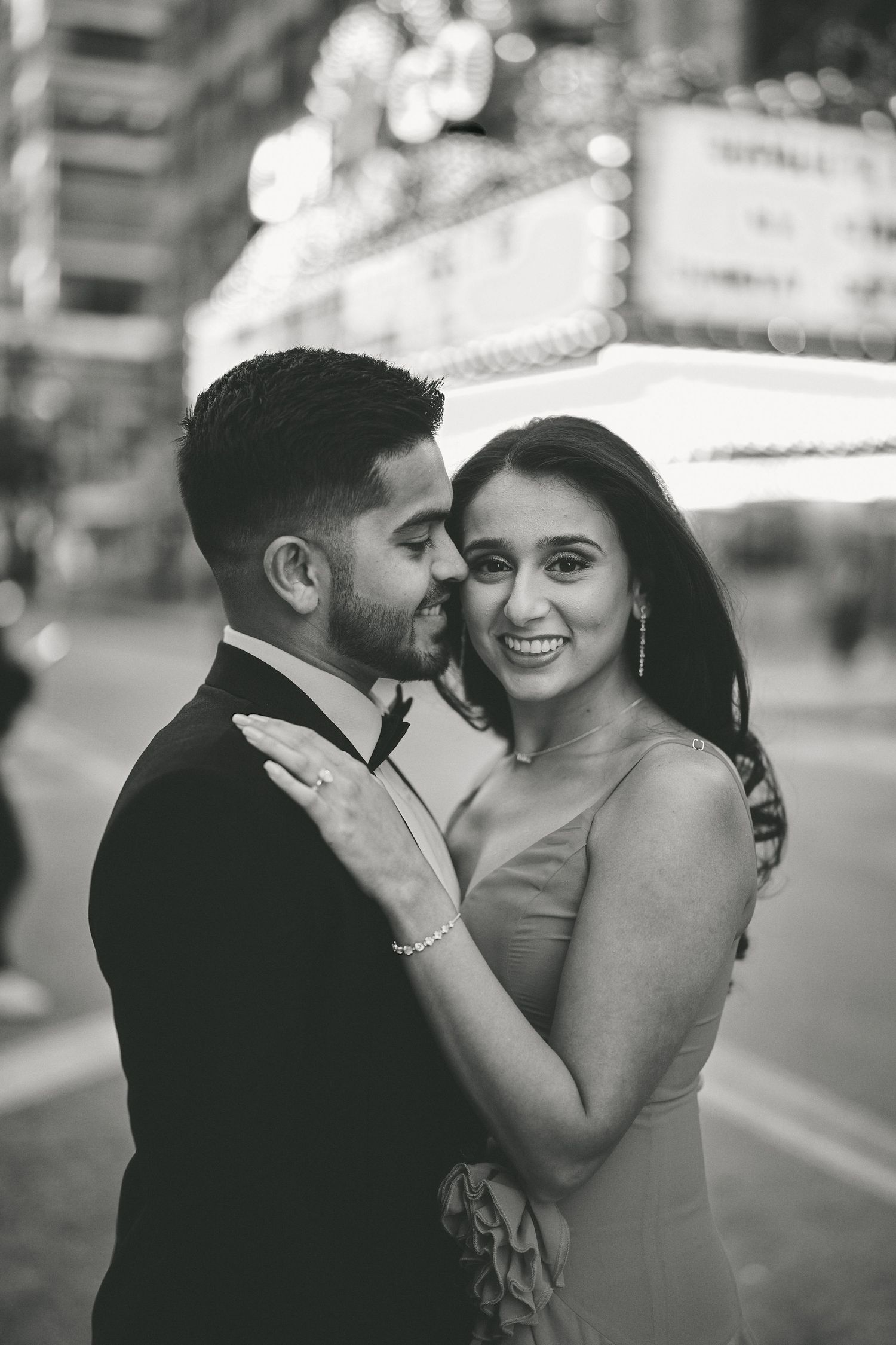 A well-dressed couple shares an intimate moment on a city street with glowing lights in the background.