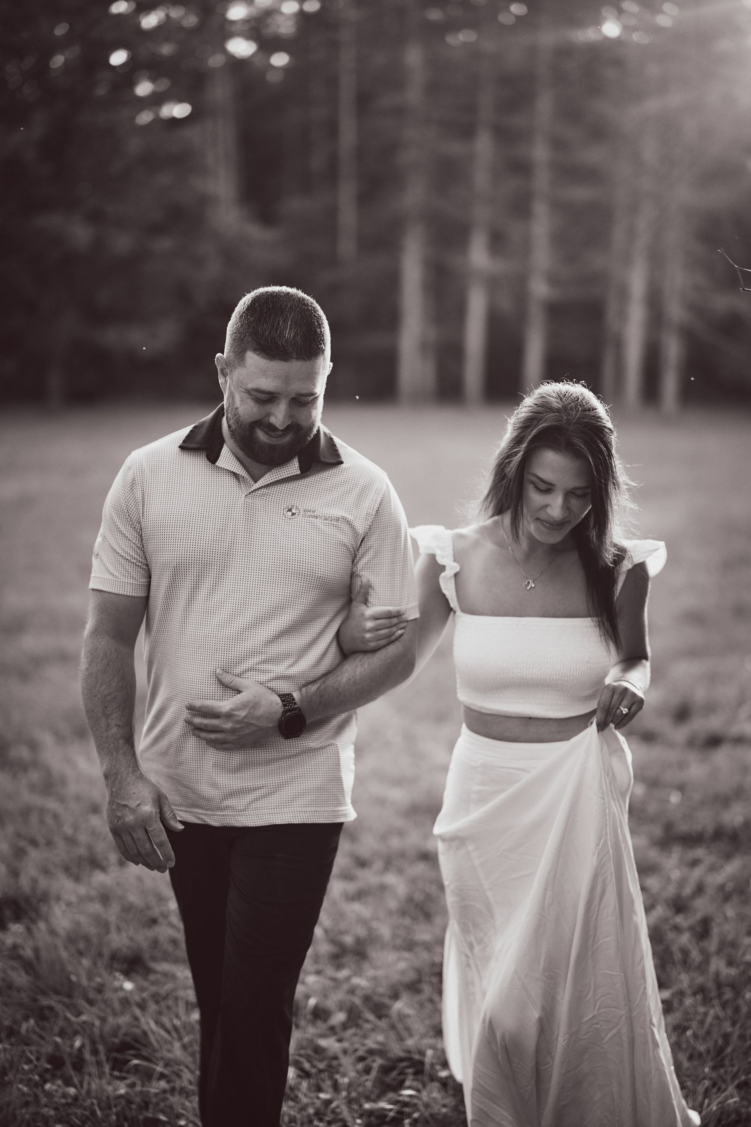 A couple in formal attire walks together through a grassy field in a black and white photograph.