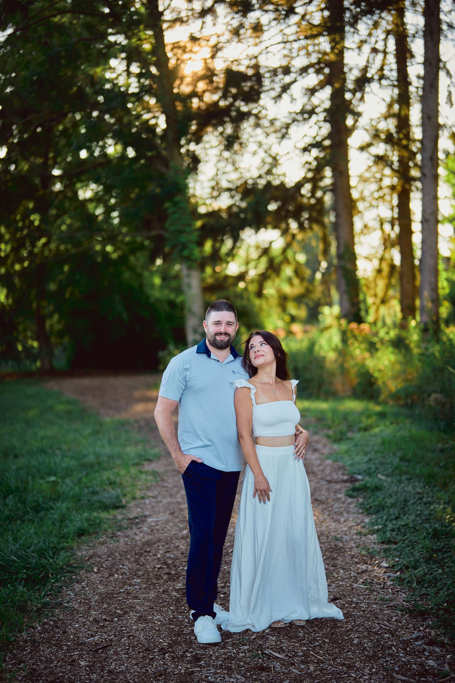 A couple stands on a sunlit forest path surrounded by towering trees during golden hour.