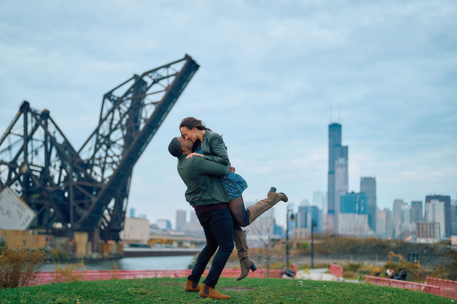 A couple embraces in front of a drawbridge and Chicago skyline along the river on an overcast day.