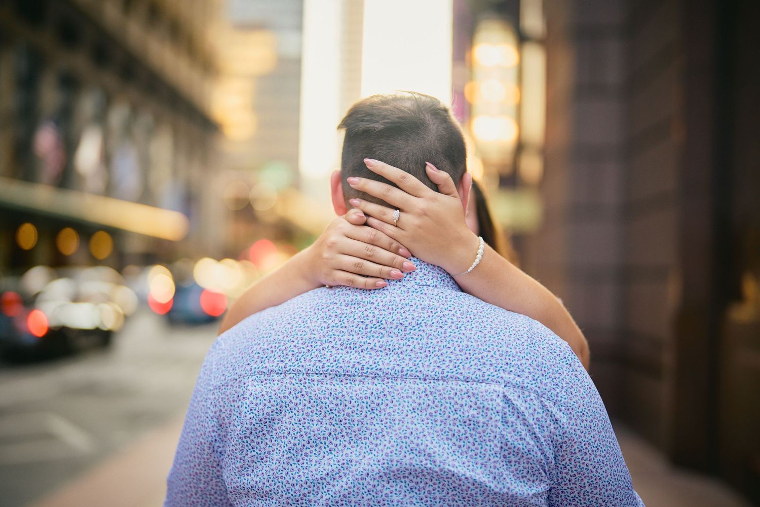 Someone hugs another person from behind with hands covering their face on an urban street with blurred background.